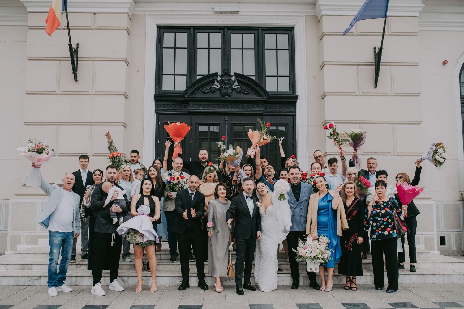 Wedding party and guests celebrate outside historic building with flowers raised in joy.
