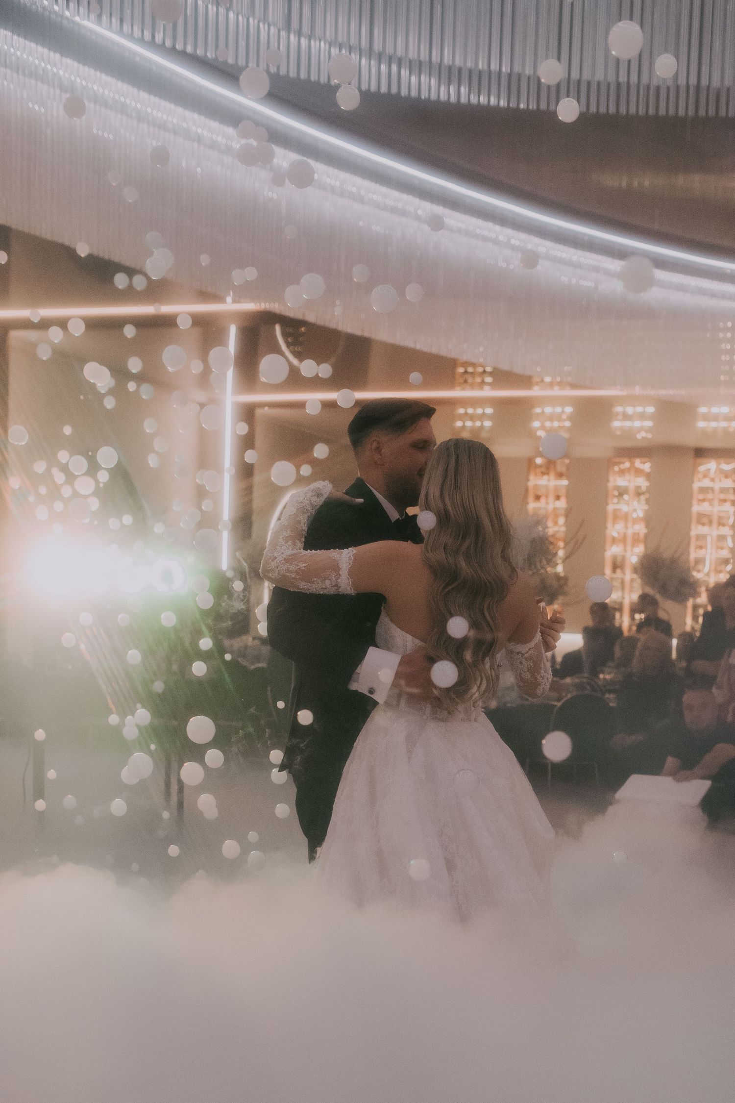 Wedding couple dancing closely amid swirling fog and sparkling bokeh lights at elegant reception venue.