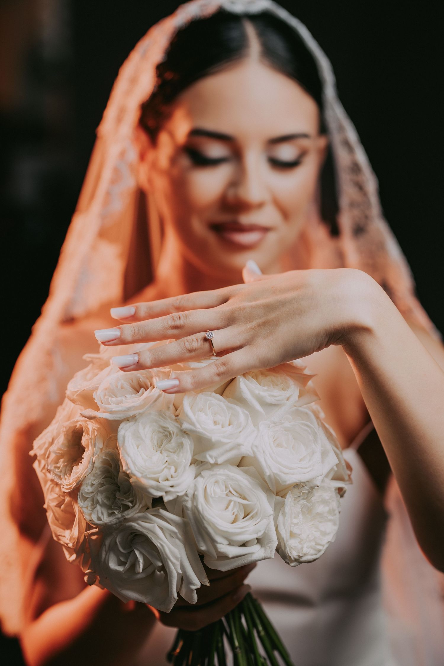 Beautiful bride wearing lace veil holding white rose bouquet with engagement ring visible on her hand.