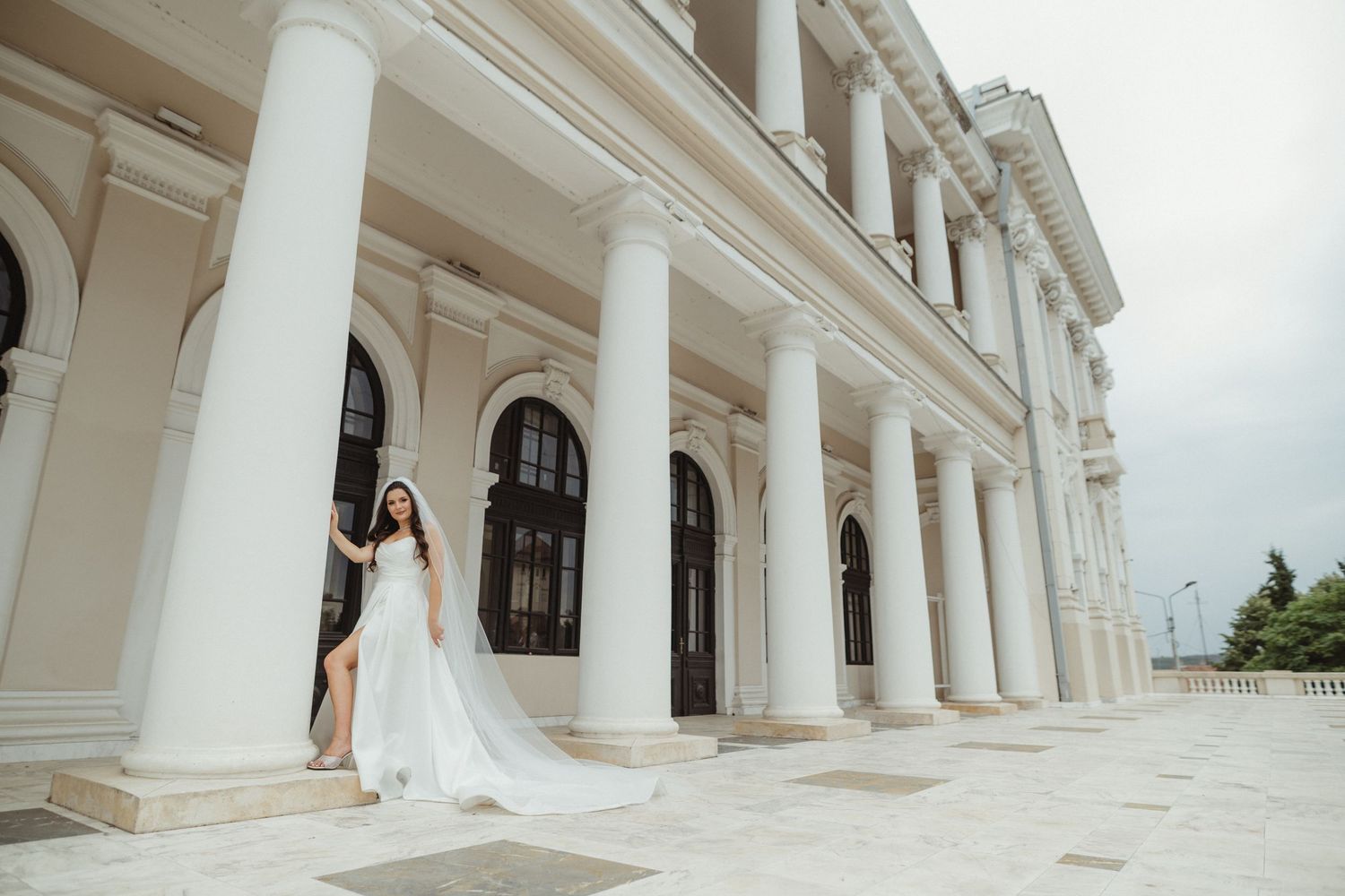 Elegant bride in a flowing white gown posing gracefully beside tall white columns of a neoclassical building.