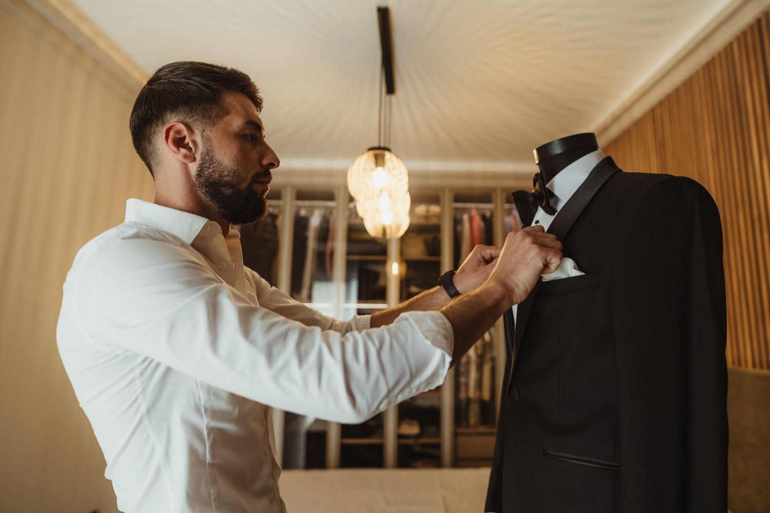 Bearded groom in white shirt adjusting boutonniere on black tuxedo displayed on a dress form.