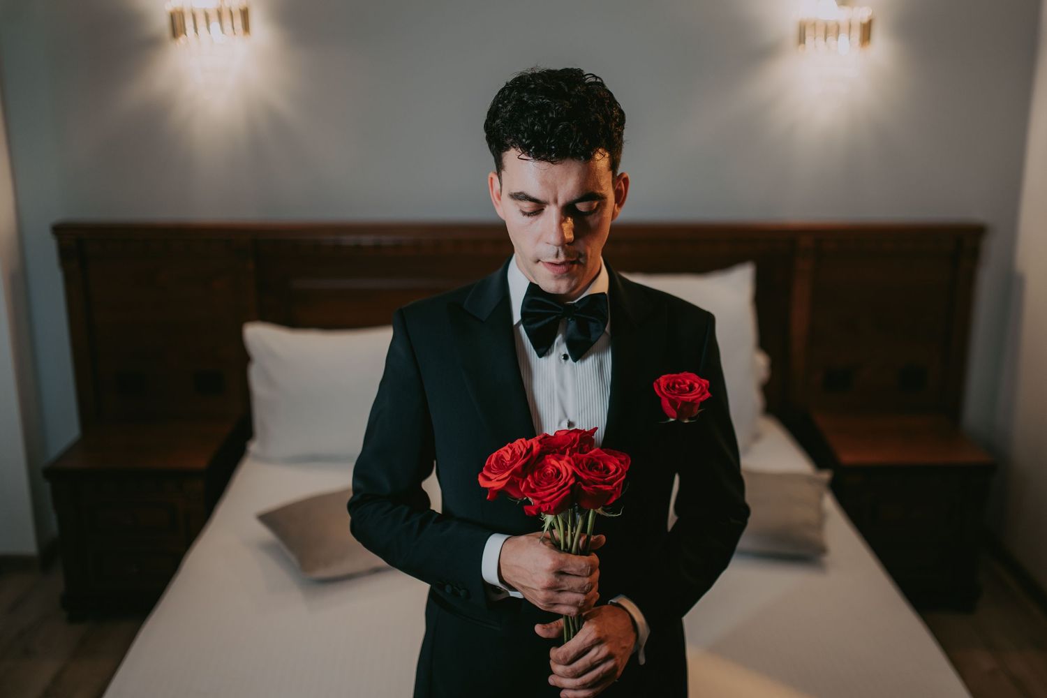 Elegant groom in black tuxedo holding red roses while standing near a hotel bed. Epicu Fotograf Drobeta-Turnu Severin