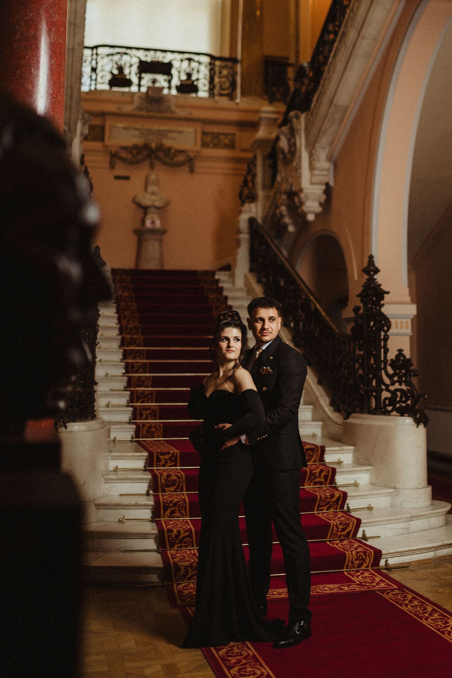 Stylish couple in black formal wear posing on grand staircase with red carpet.