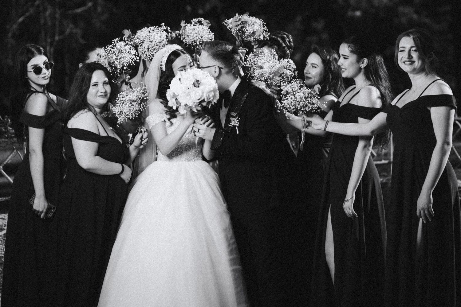 Black and white photo of bride and groom kissing surrounded by bridesmaids holding flower bouquets.