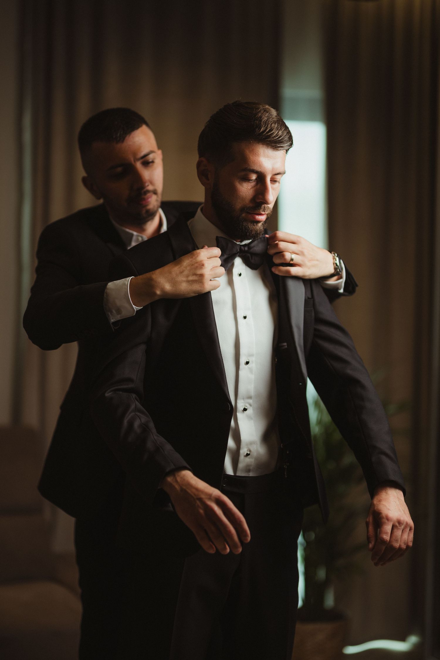 Best man helping groom adjust black bow tie on tuxedo while preparing for wedding ceremony.
