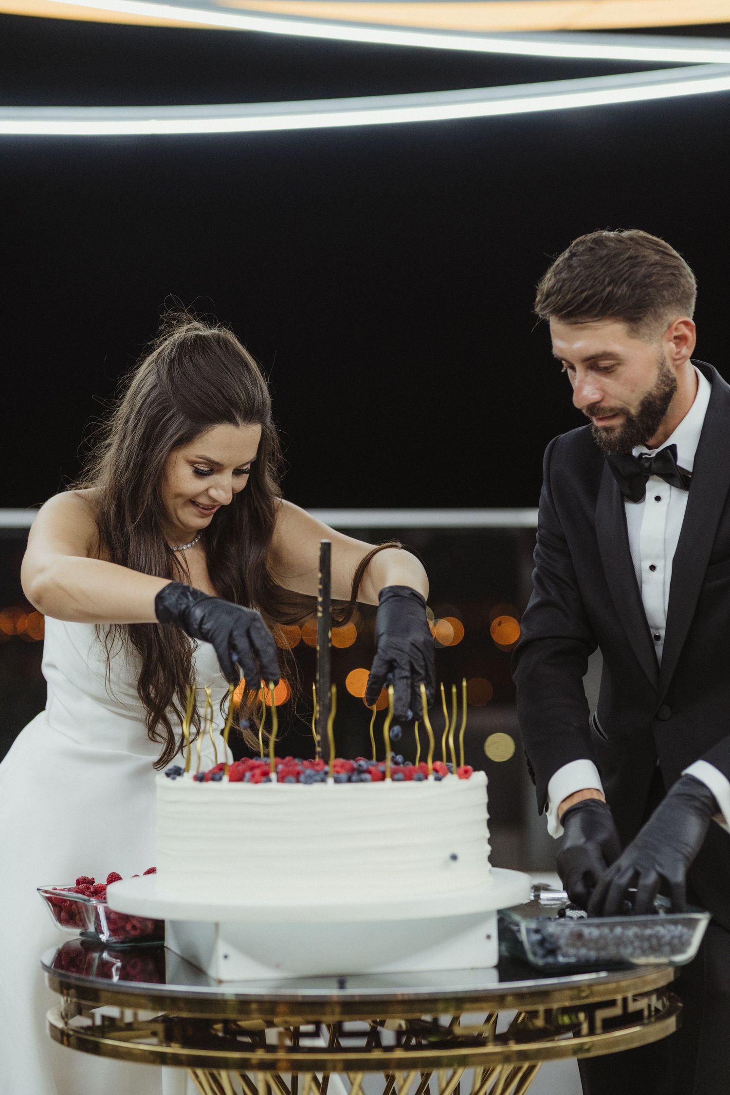 Smiling bride and groom in black gloves cutting decorated white tiered wedding cake at reception. Epicu Fotograf Drobeta-Turnu Severin