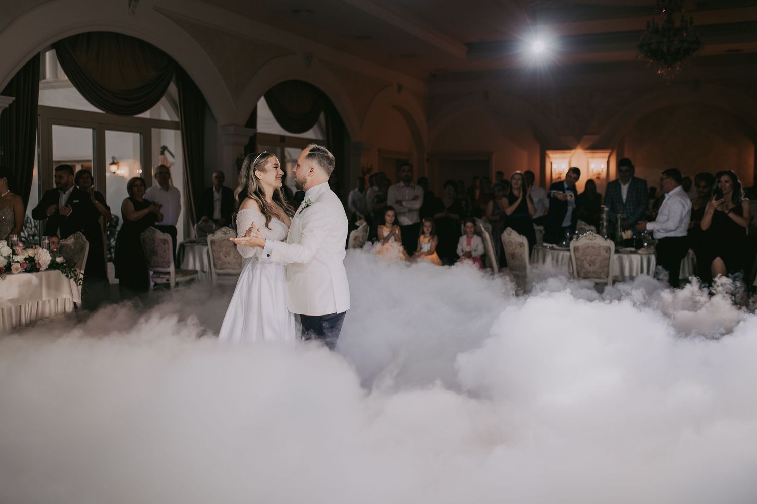 Newlyweds dancing together on a cloud of fog in an elegant chandelier-lit wedding reception hall. Epicu Fotograf Drobeta-Turnu Severin