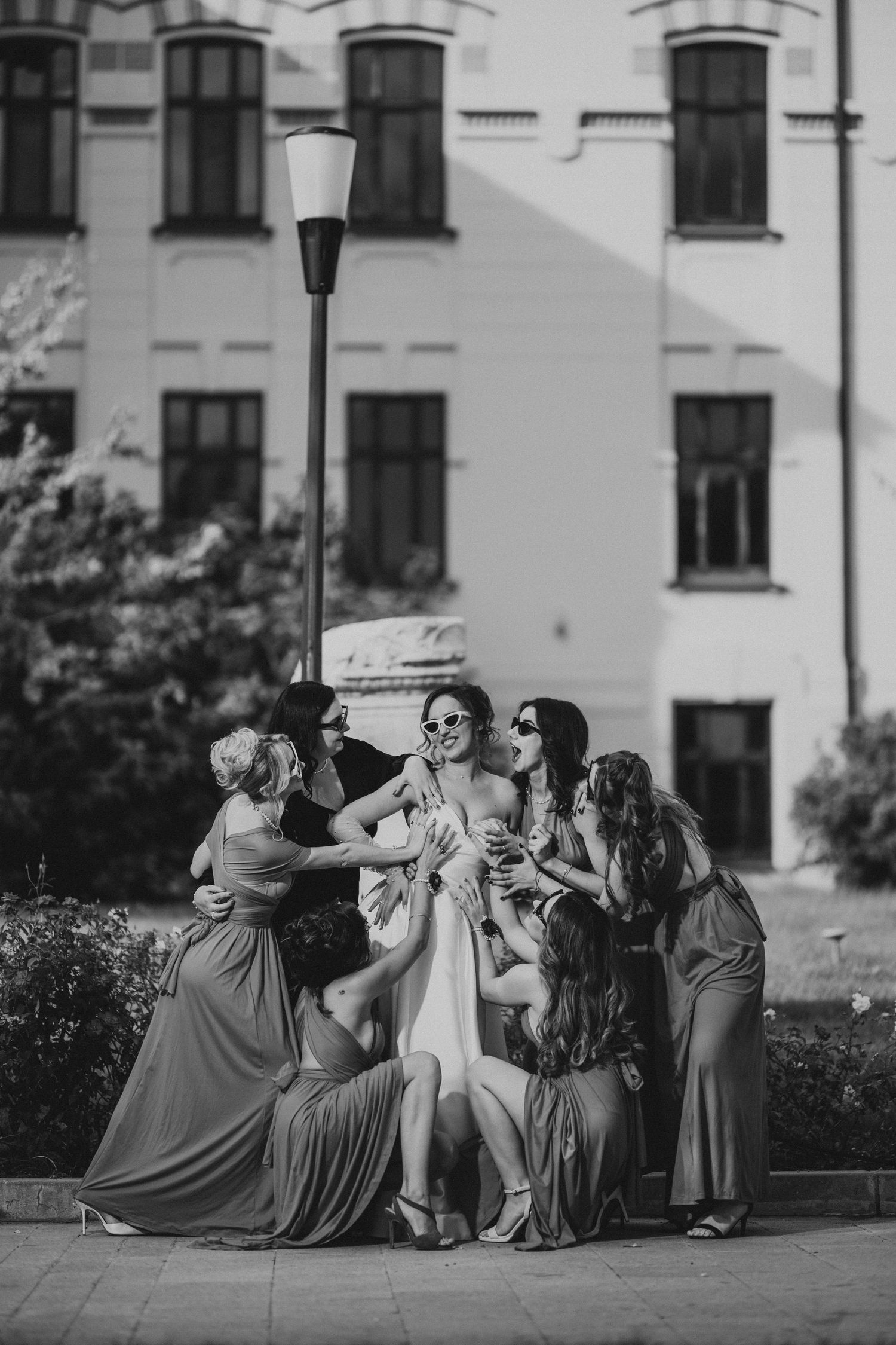 Black and white photo of bride surrounded by bridesmaids embracing near a building. Epicu Fotograf Drobeta-Turnu Severin