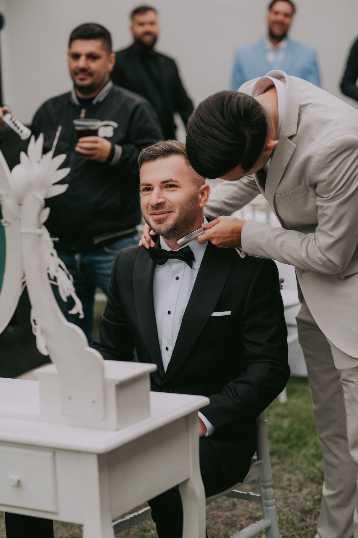 Groomsman helping groom with bow tie while seated at white vanity table during outdoor wedding prep.