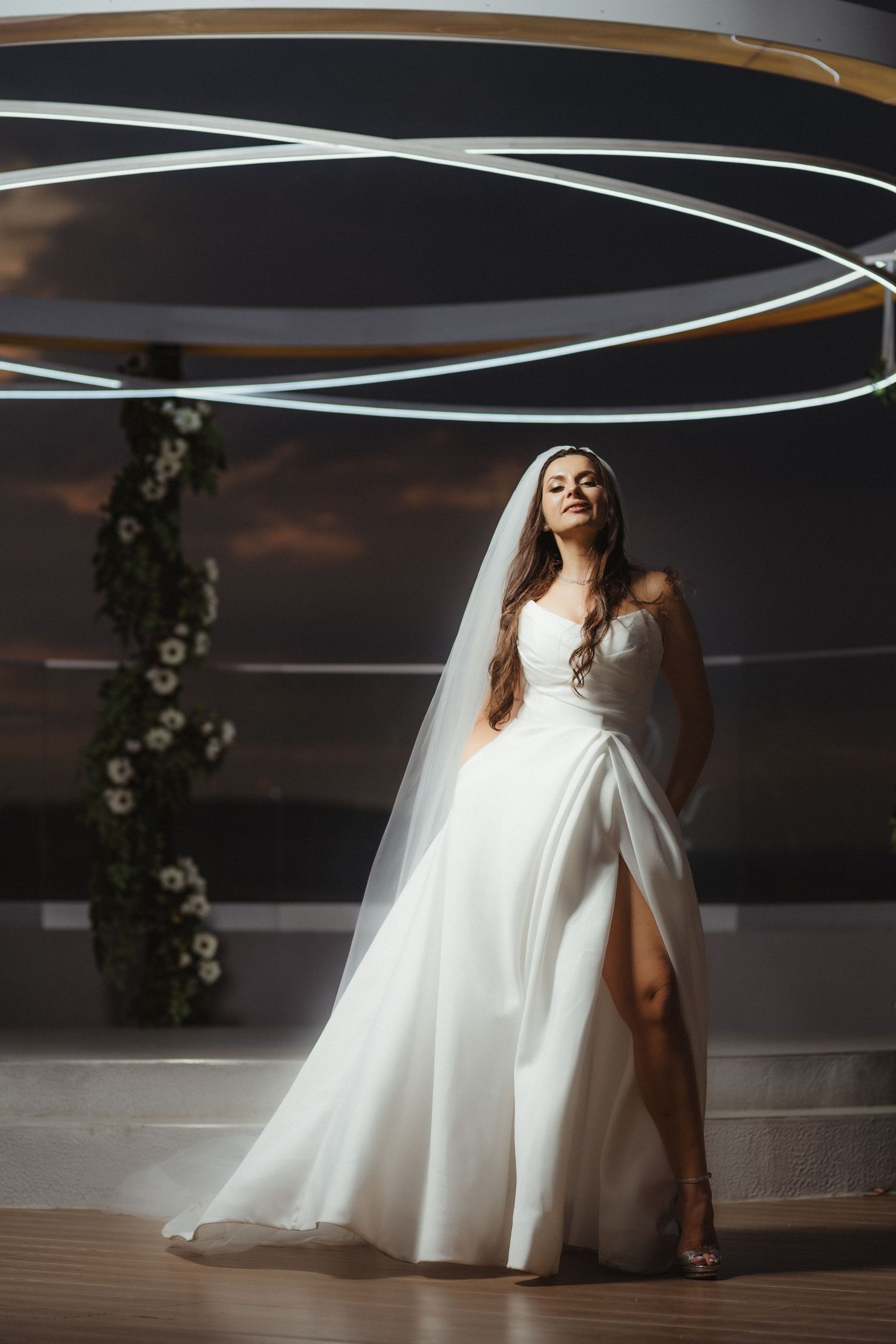 Bride in strapless white gown with thigh slit poses gracefully under circular neon lights at rooftop wedding venue.