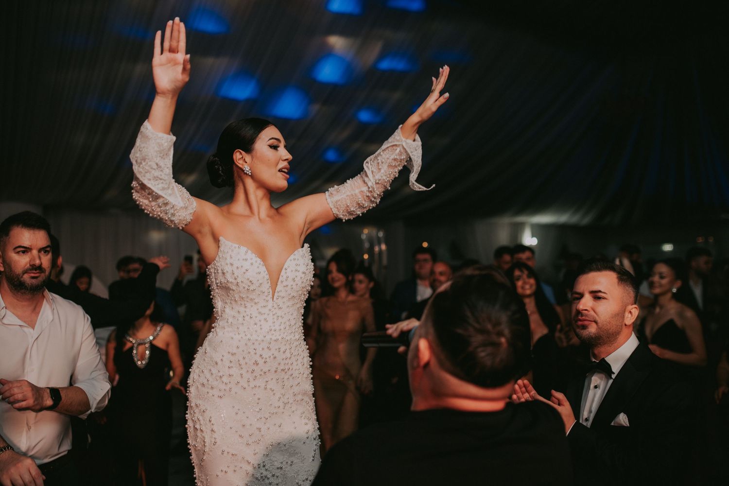 Bride in pearl-encrusted gown raises her arms joyfully while being celebrated by guests on the dance floor.