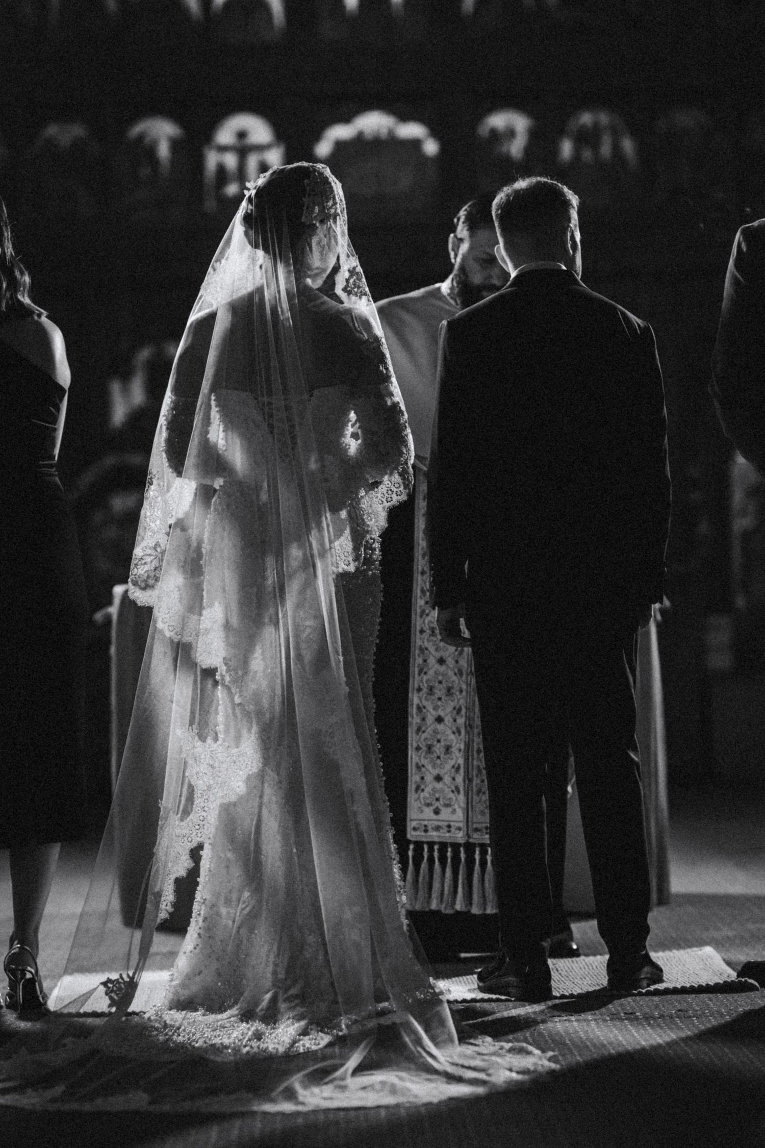 Black and white photo of bride in lace veil and groom standing before priest at church altar.