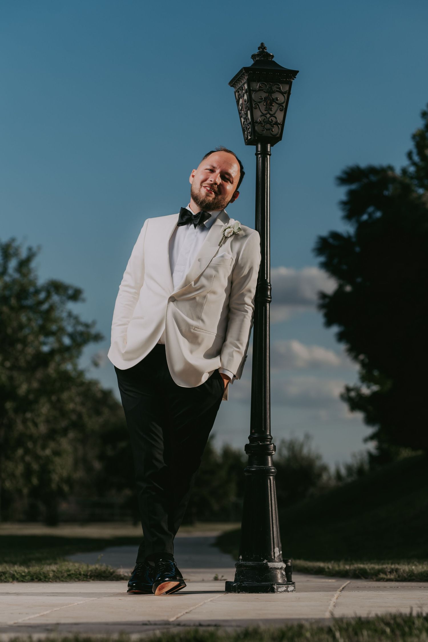 Stylish groom in white dinner jacket leans against ornate black lamppost under blue sky. Epicu Fotograf Drobeta-Turnu Severin