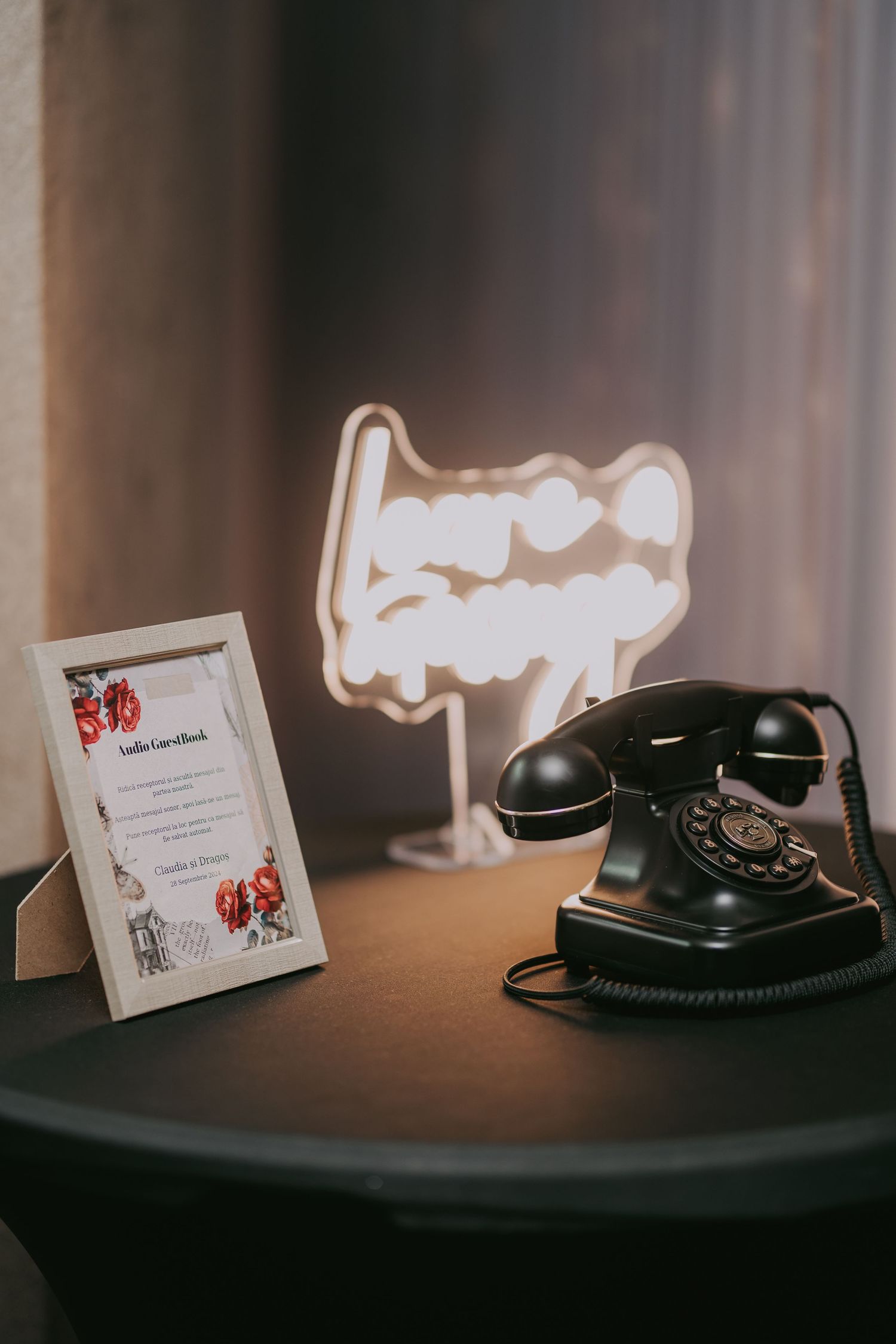 Vintage black rotary telephone beside a glowing love neon sign and framed wedding guest book card on a dark table. Epicu Fotograf Drobeta
