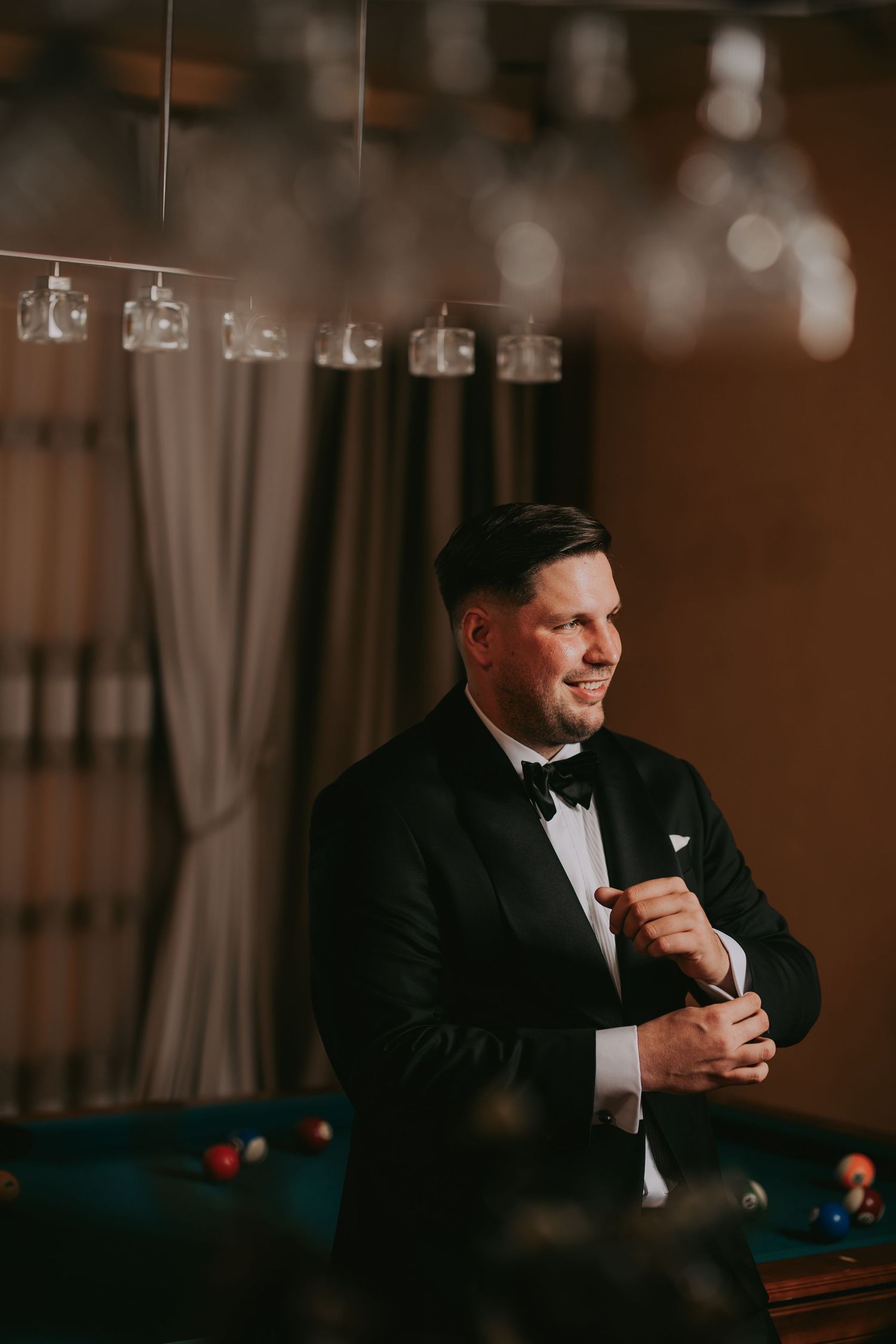 Elegant man in black tuxedo with bow tie stands confidently near a billiard table indoors.