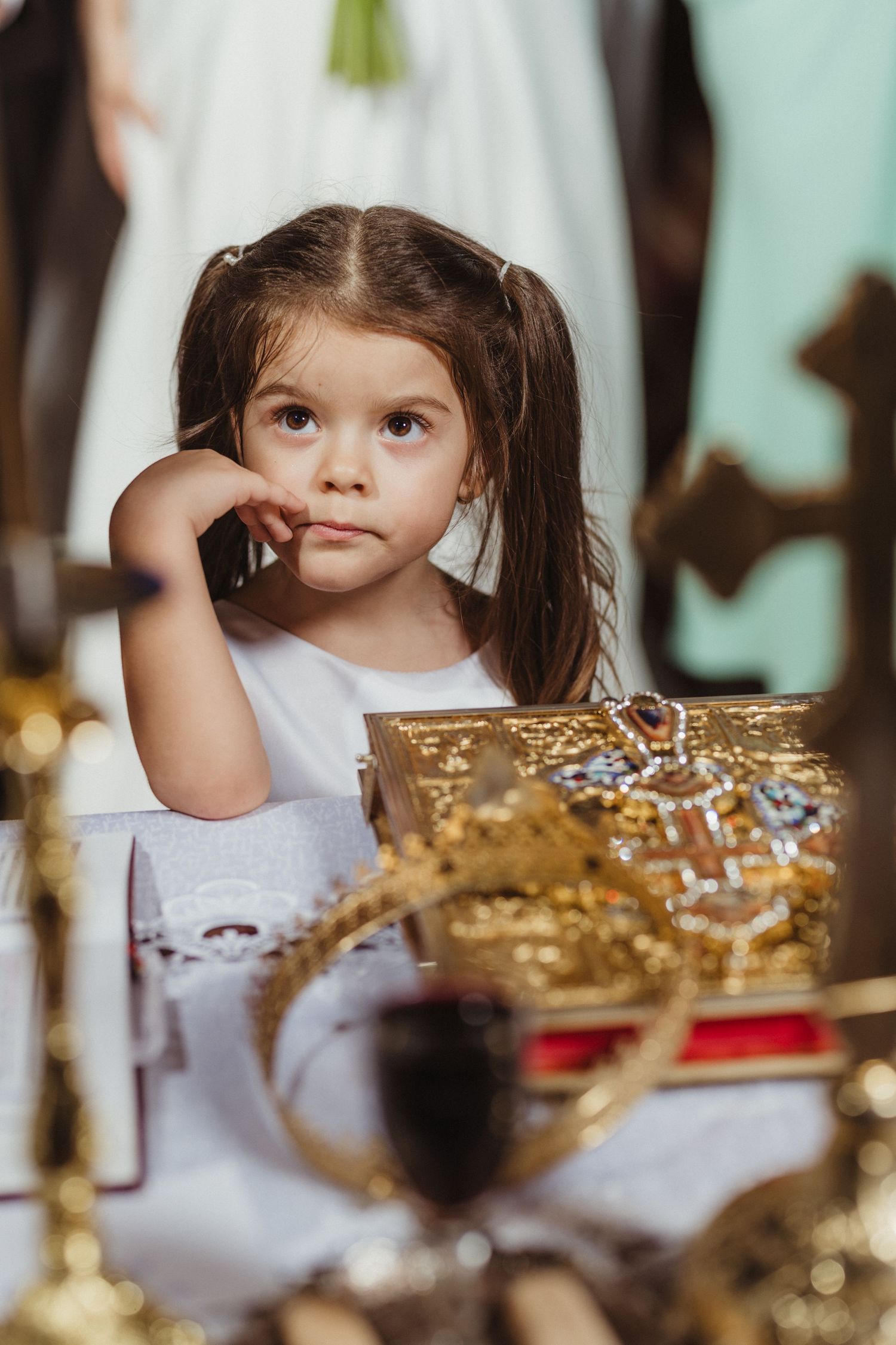 Adorable little girl in white dress resting her chin on her hand near golden ceremonial items at a wedding.