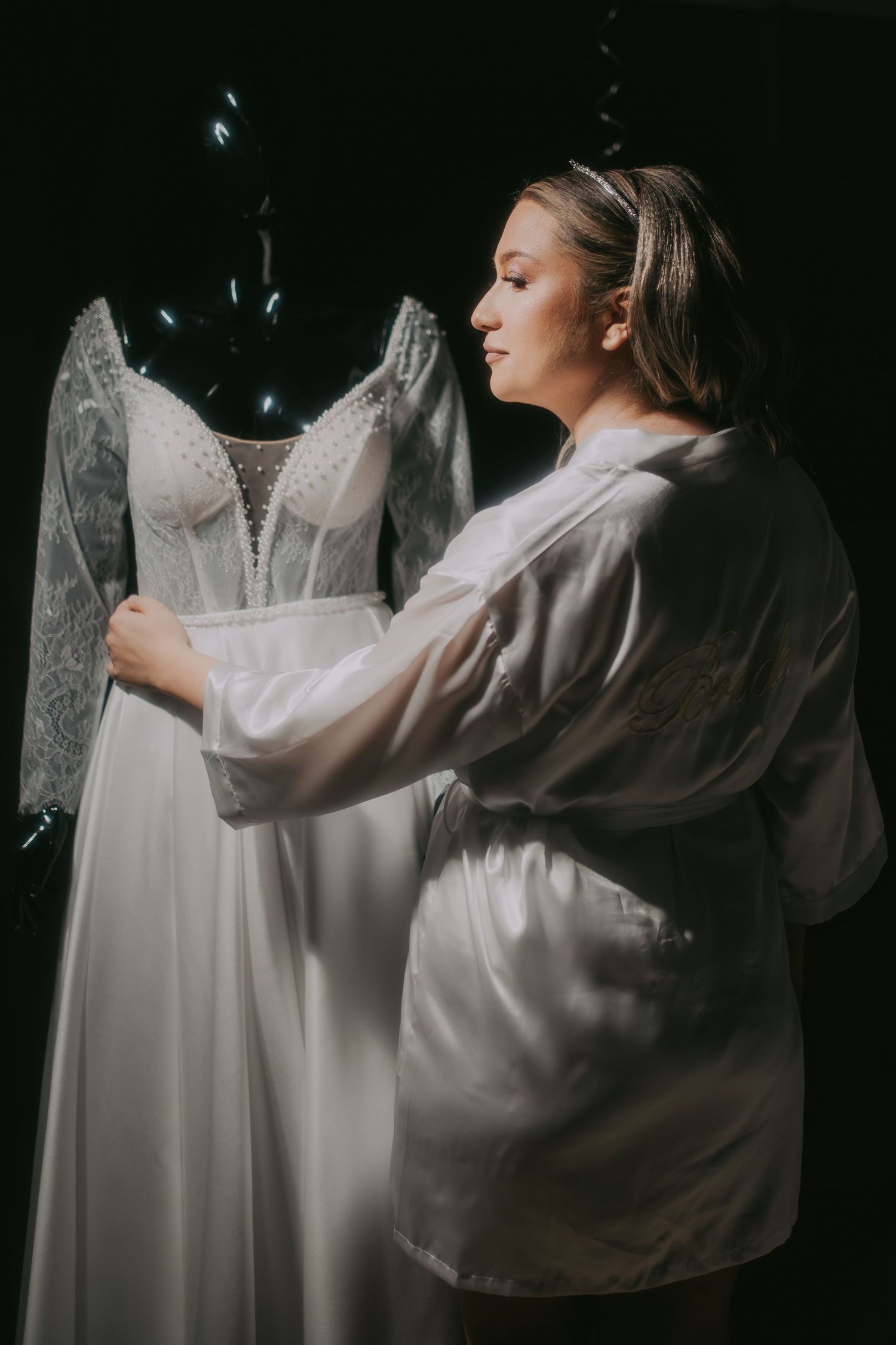 Bride in white robe admiring her lace wedding gown on a mannequin against a dark background. Epicu Fotograf Drobeta-Turnu Severin