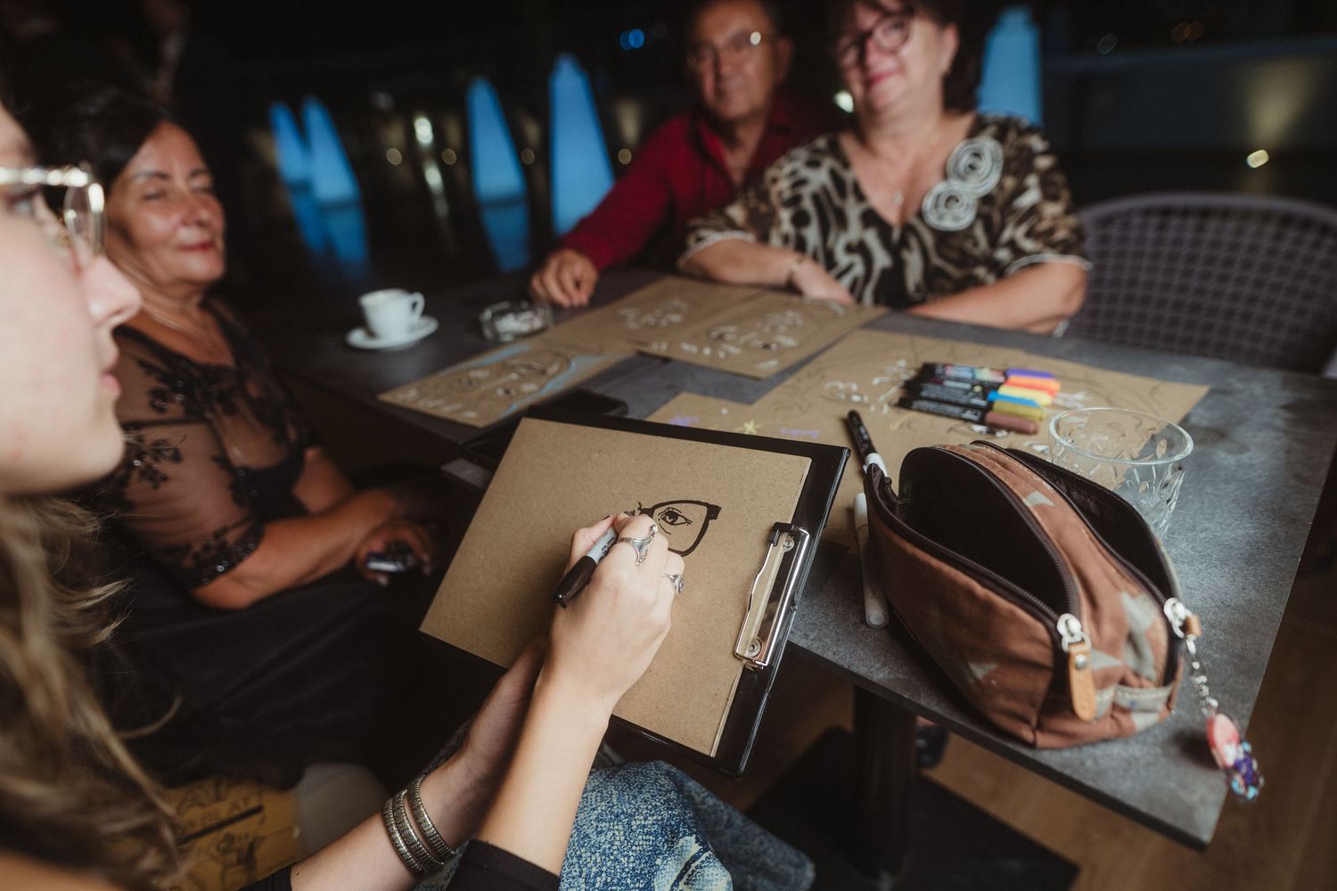 Women gather around table engaged in creative drawing activity at casual evening event.