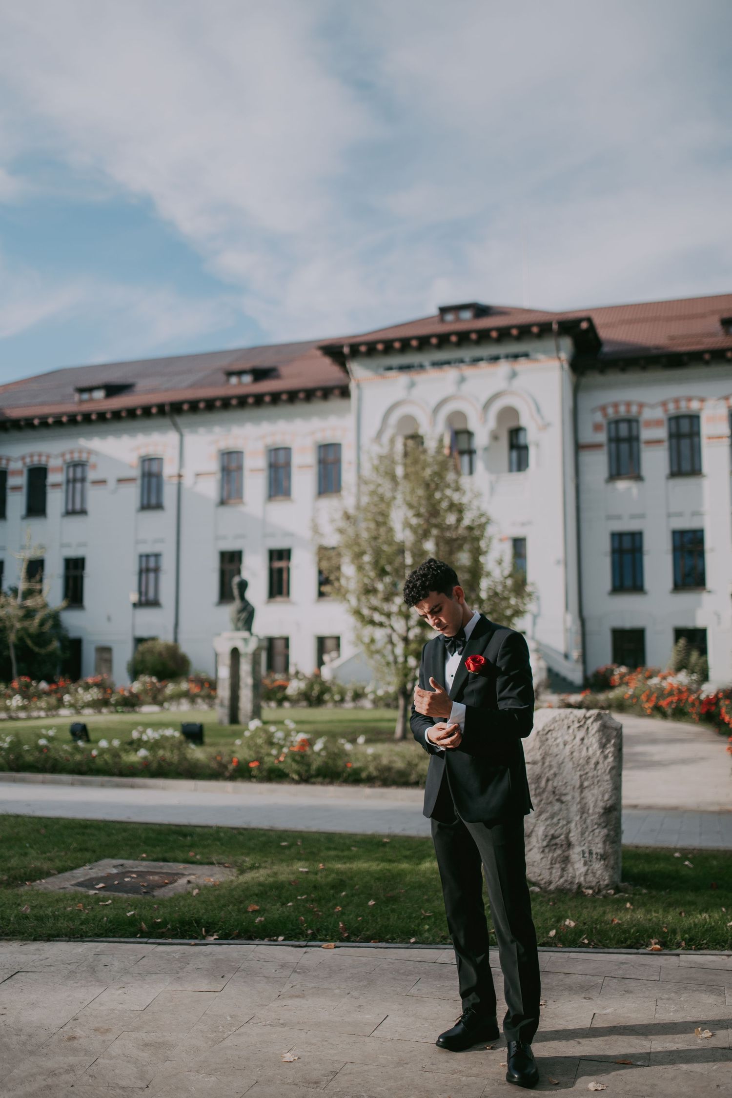 Handsome groom in black suit standing before a grand white building in a blooming garden. Epicu Fotograf Drobeta-Turnu Severin