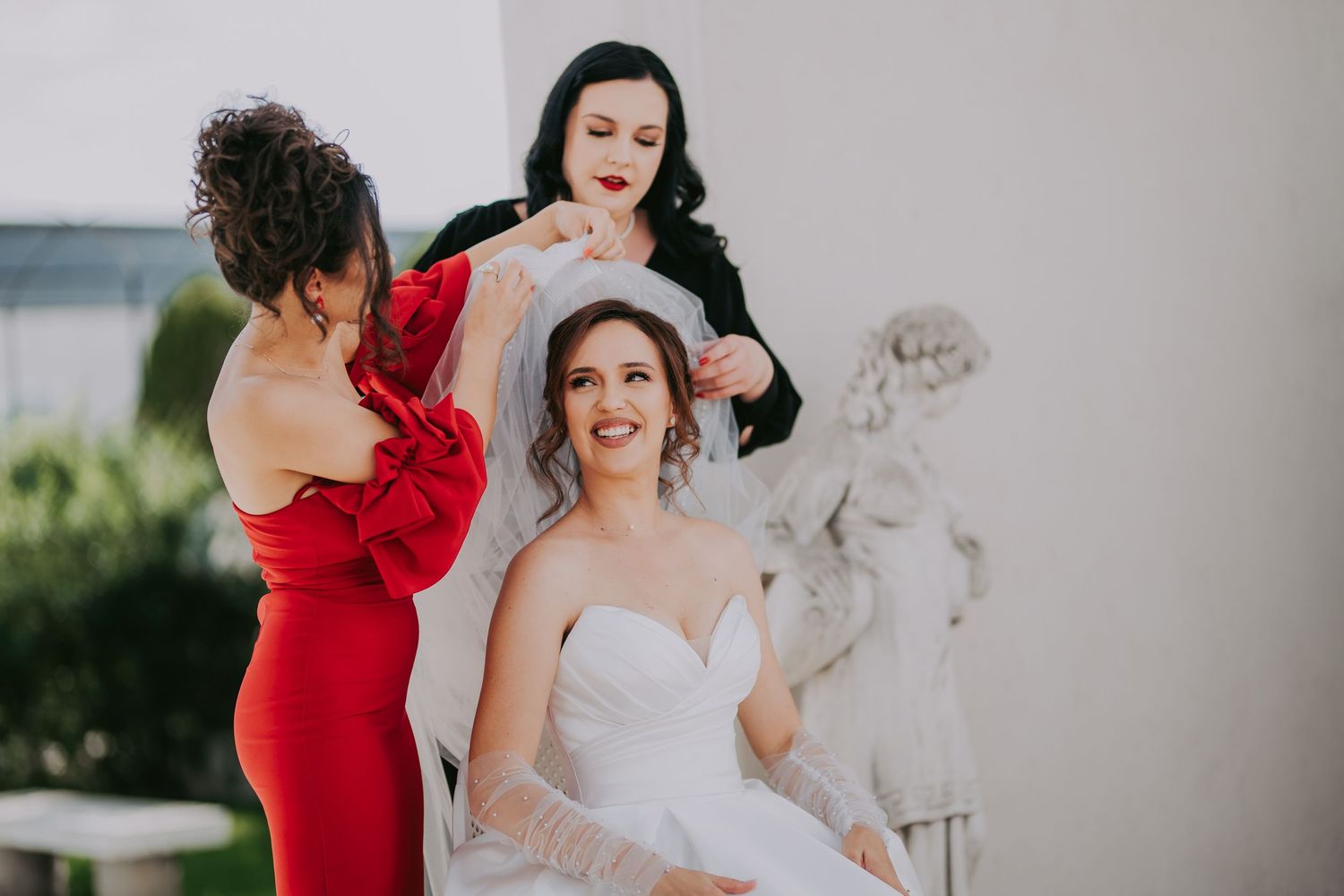 Bride in white gown being helped with her veil by two women in red and black dresses. Epicu Fotograf Drobeta-Turnu Severin