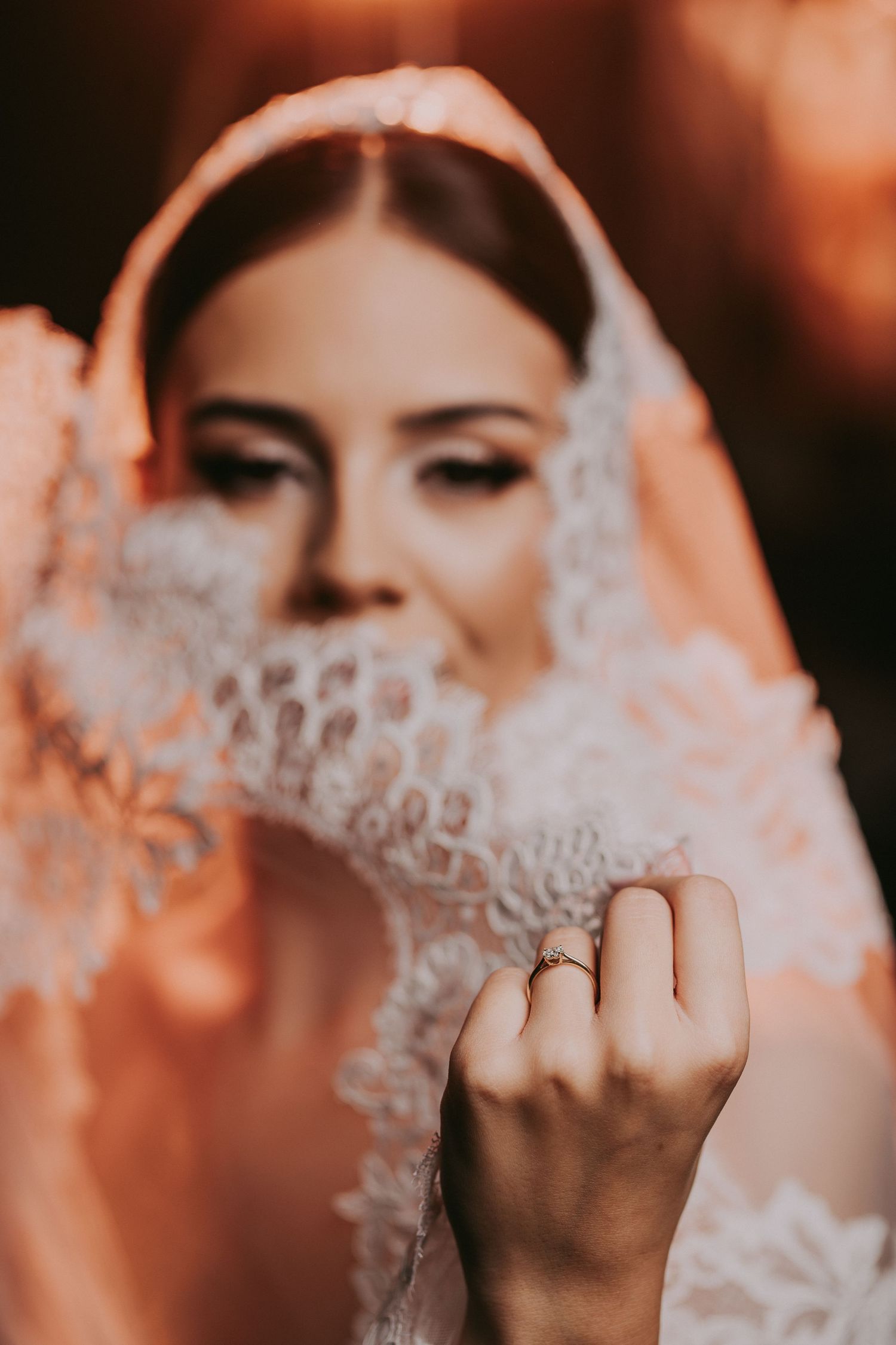 Bride in lace veil showing diamond engagement ring on her finger in a soft warm-toned wedding portrait.