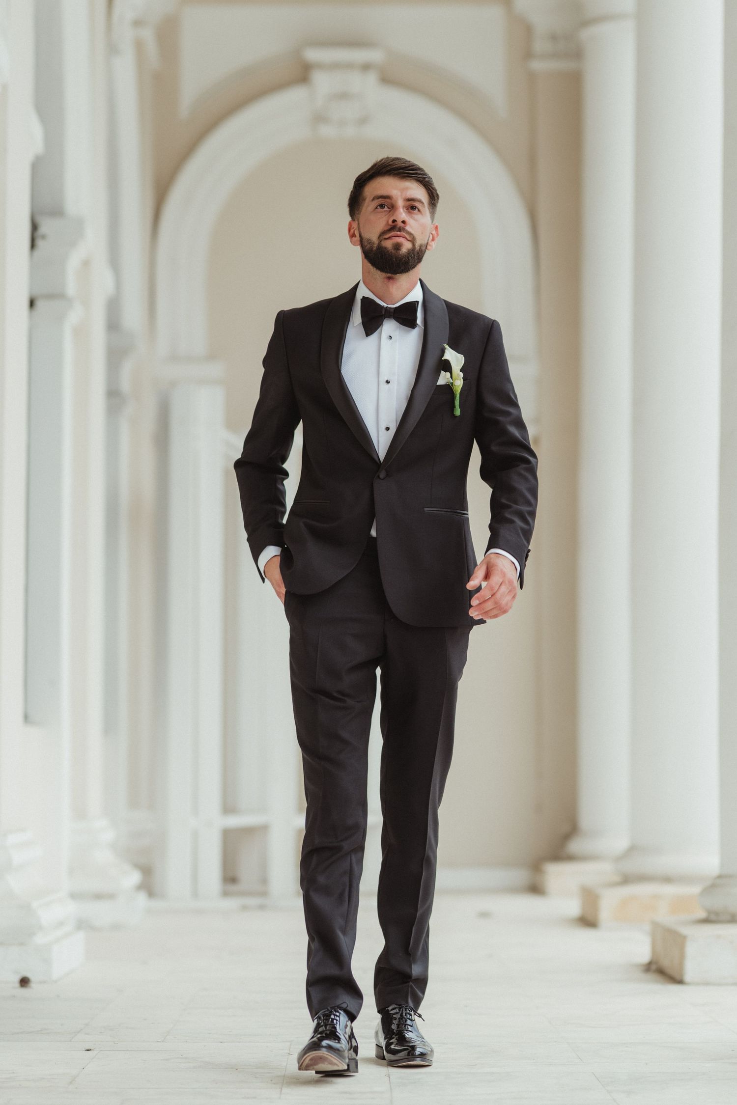 Handsome bearded groom in black tuxedo with boutonniere walking confidently through arched corridor.
