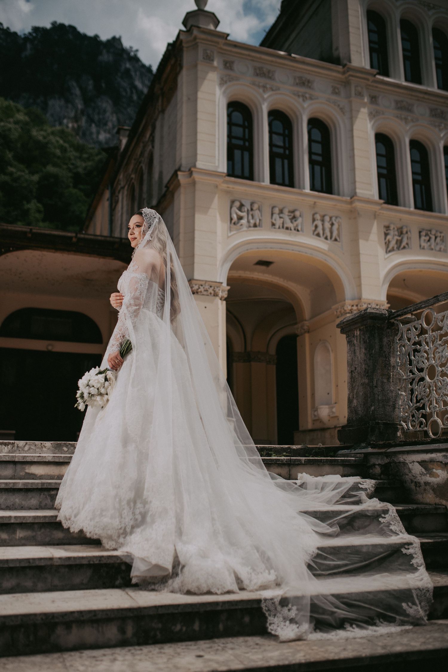Stunning bride in lace gown and flowing cathedral veil poses on steps of historic building.