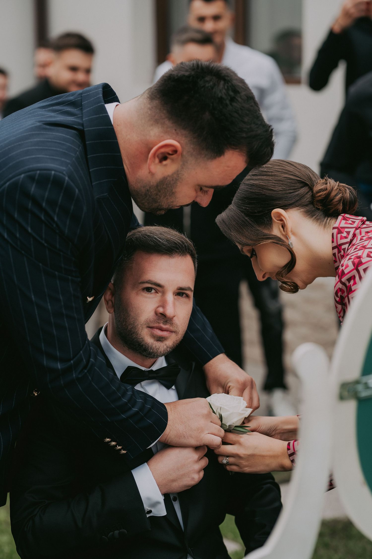 Two groomsmen pinning white boutonniere on groom's lapel during outdoor wedding preparation ceremony.