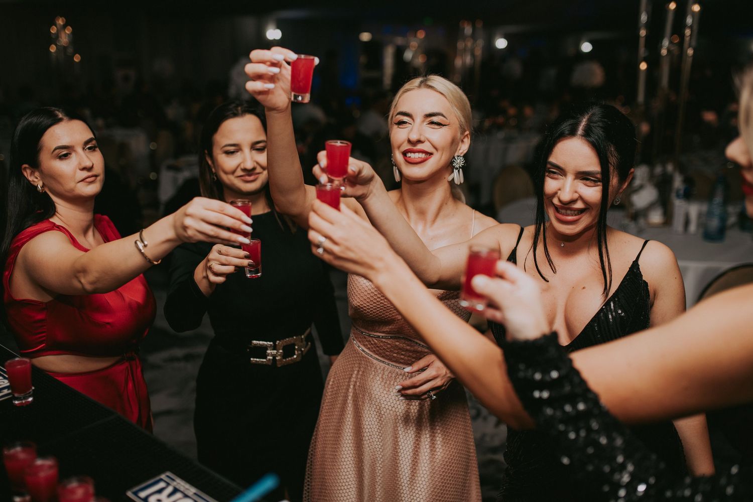 Group of elegant women in formal dresses toasting with red shot glasses at a party celebration.