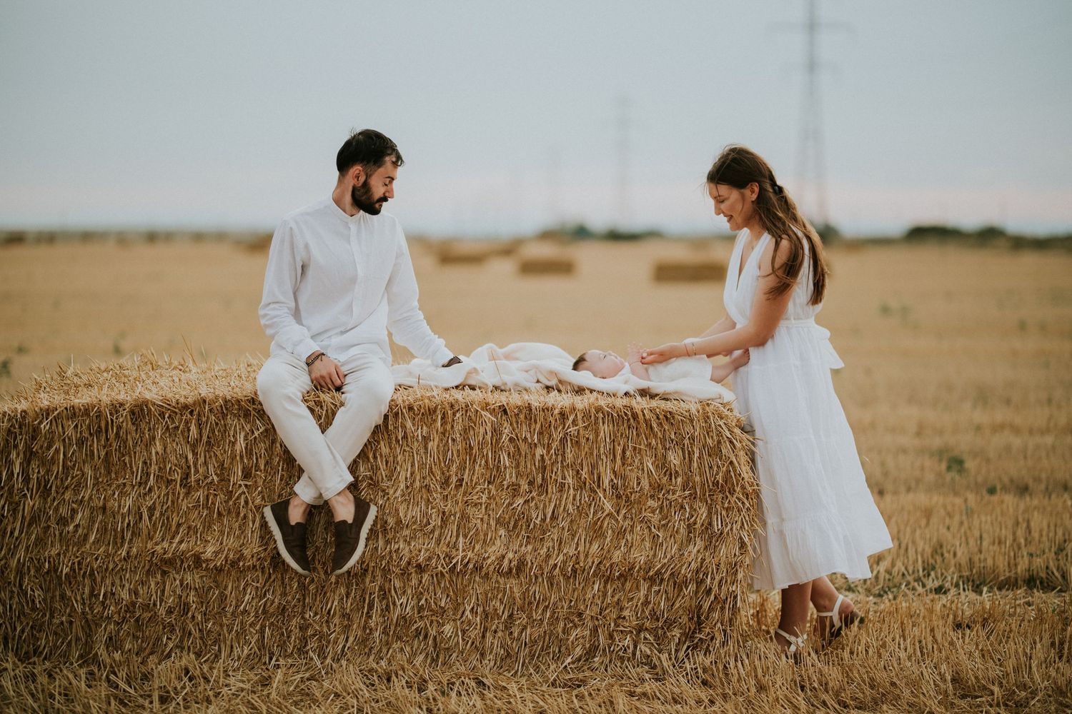 Couple in white outfits sitting on a hay bale in a golden wheat field at sunset.