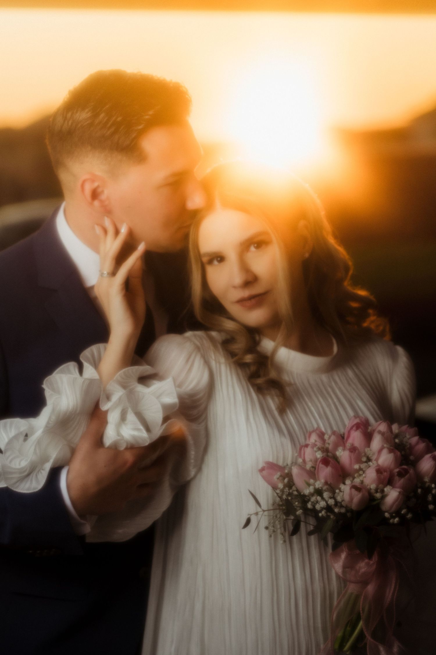 Romantic couple embraces at golden sunset, bride holding pink tulip bouquet in elegant white dress.