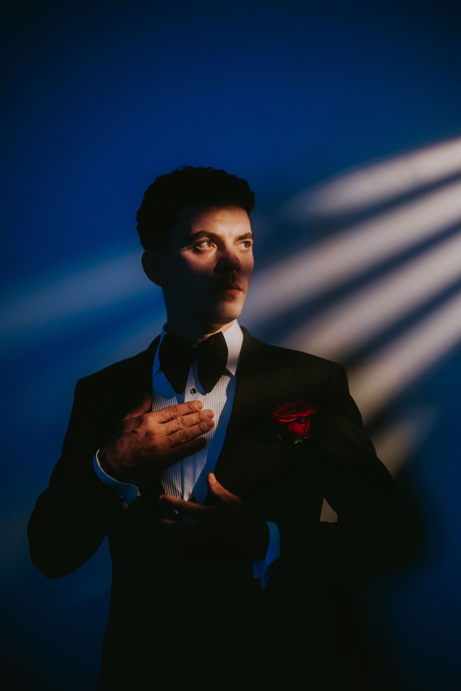 Elegant groom in black tuxedo with red pocket square posing against blue backdrop.
