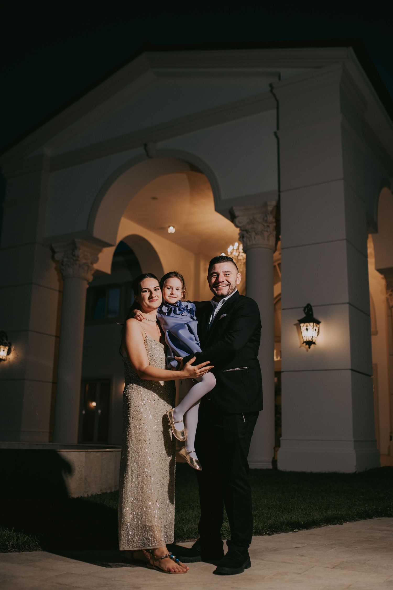 Smiling family posing at night outside illuminated venue, parents holding young child in formal attire. Epicu Fotograf Drobeta-Turnu Severin
