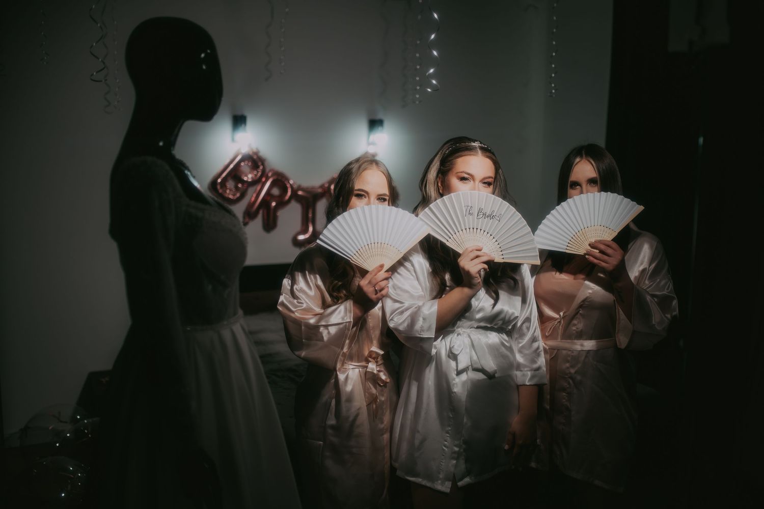 Bridesmaids in white robes holding decorative fans near a wedding dress on a mannequin in dim lighting. Epicu Fotograf Drobeta-Turnu Severin