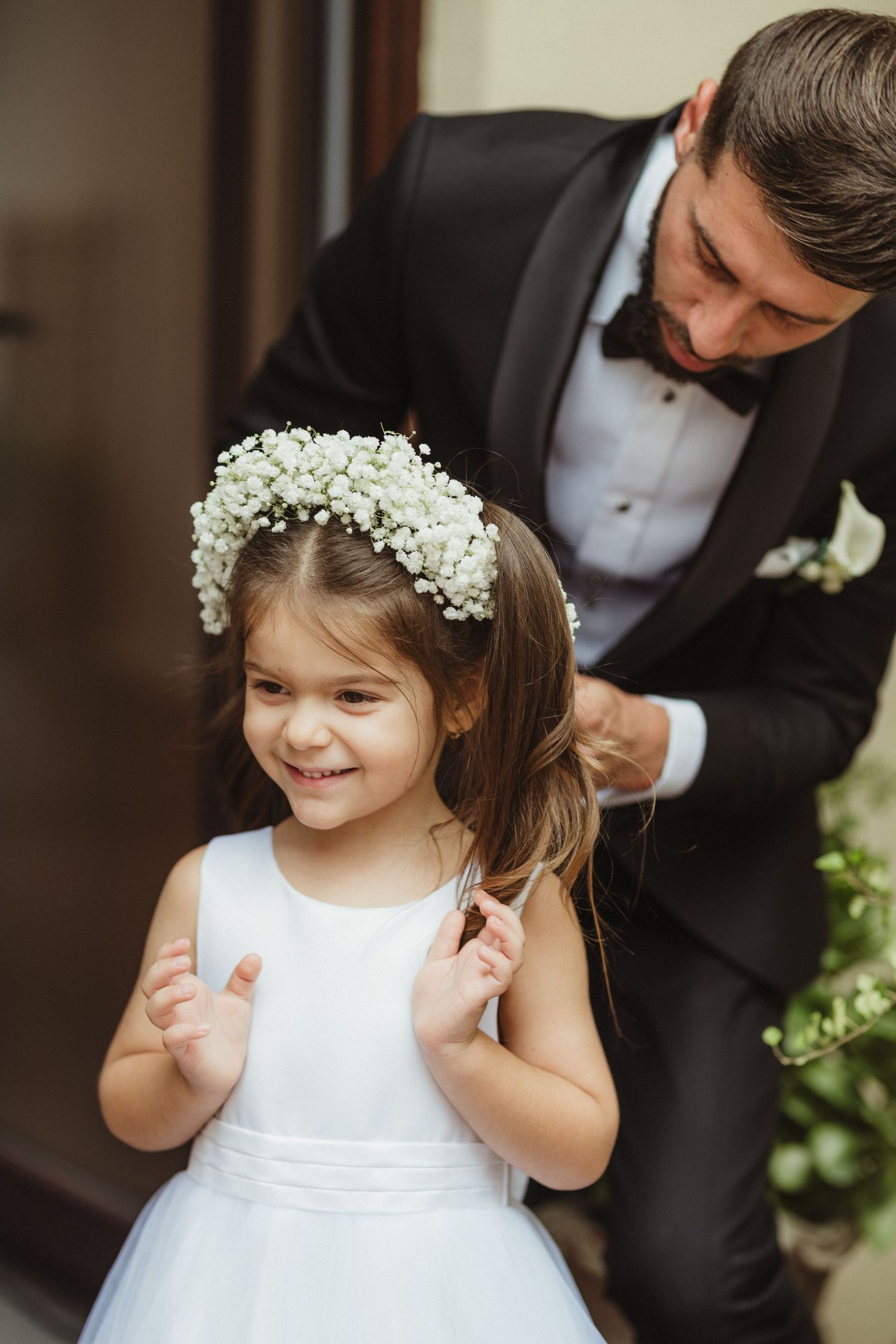 Smiling flower girl in white dress with baby's breath crown, groom in black tuxedo behind her.