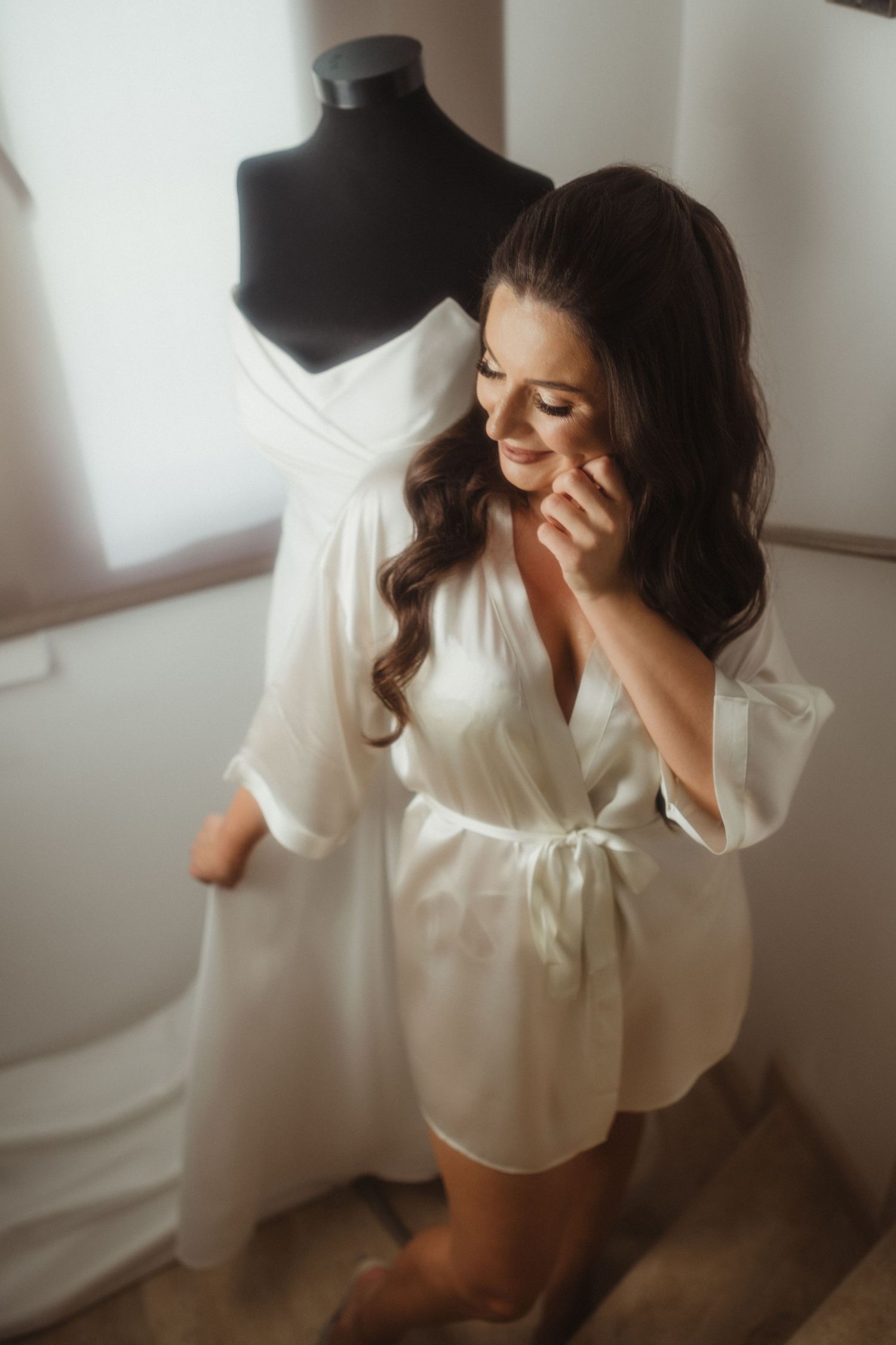 Smiling bride in white silk robe touching face while standing near wedding dress on mannequin.