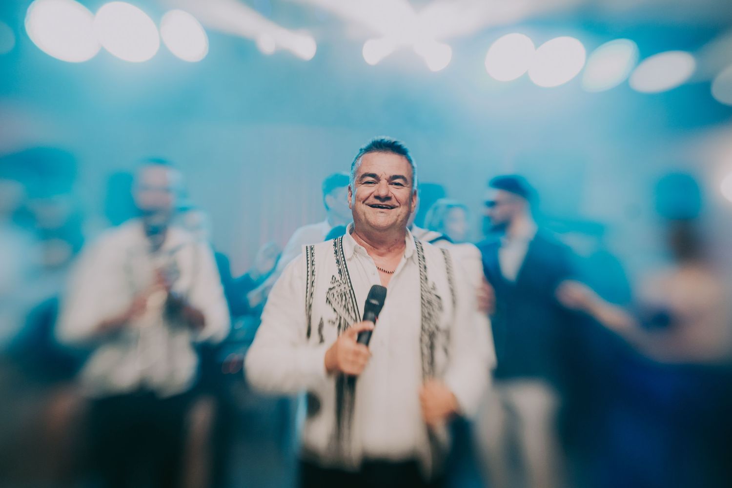 Smiling man holding microphone performing in traditional embroidered shirt at wedding reception. Epicu Fotograf Drobeta-Turnu Severin