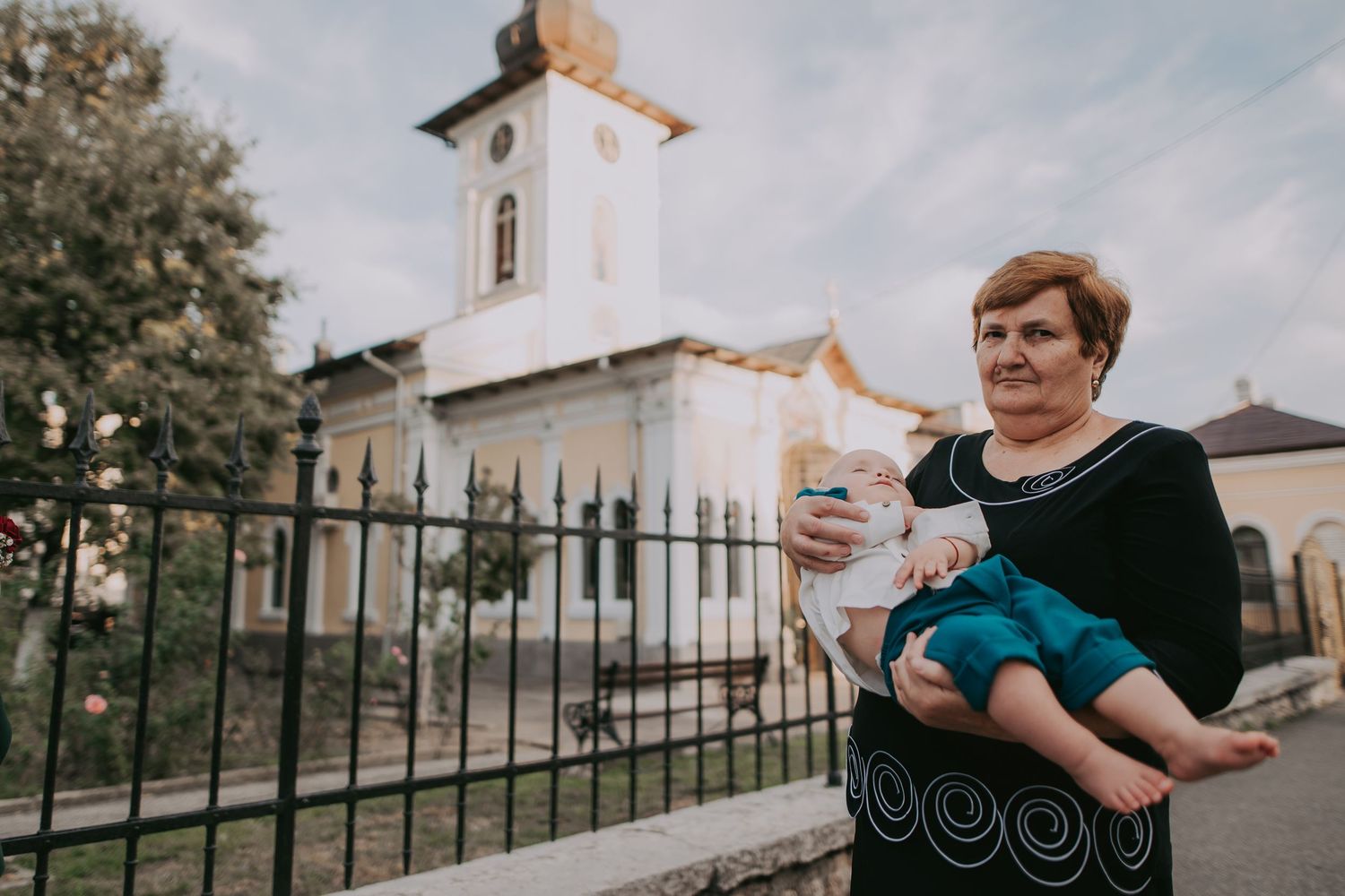 Elderly woman holding baby outside white Orthodox church with golden dome. Epicu Fotograf Drobeta-Turnu Severin