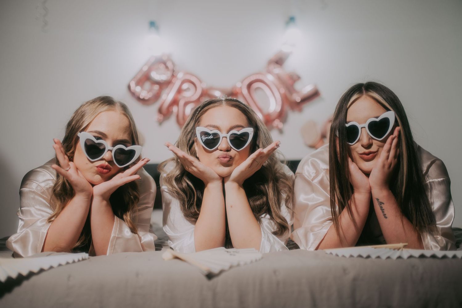 Three bridesmaids wearing heart-shaped sunglasses posing on a bed with a BRIDE balloon sign backdrop. Epicu Fotograf Drobeta-Turnu Severin