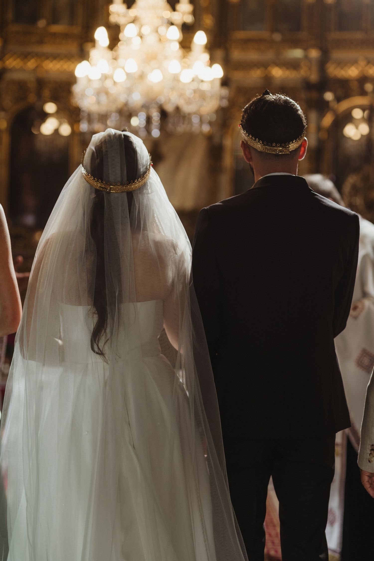 Bride and groom seen from behind wearing golden crowns before a glowing chandelier in an Orthodox church.