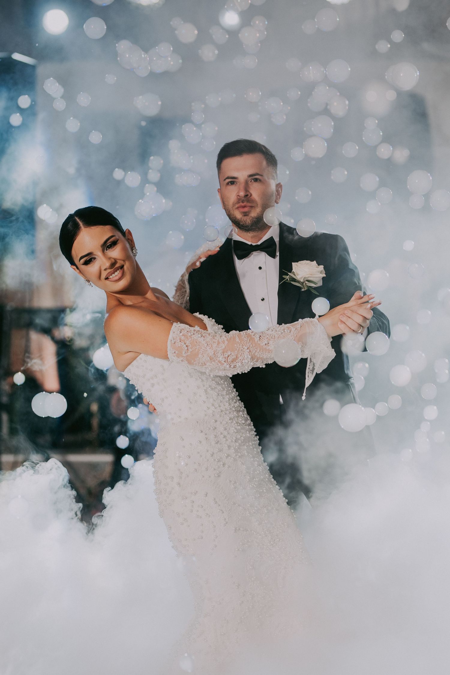 Smiling bride and groom dance together enveloped in white fog with glowing bokeh lights at their wedding.