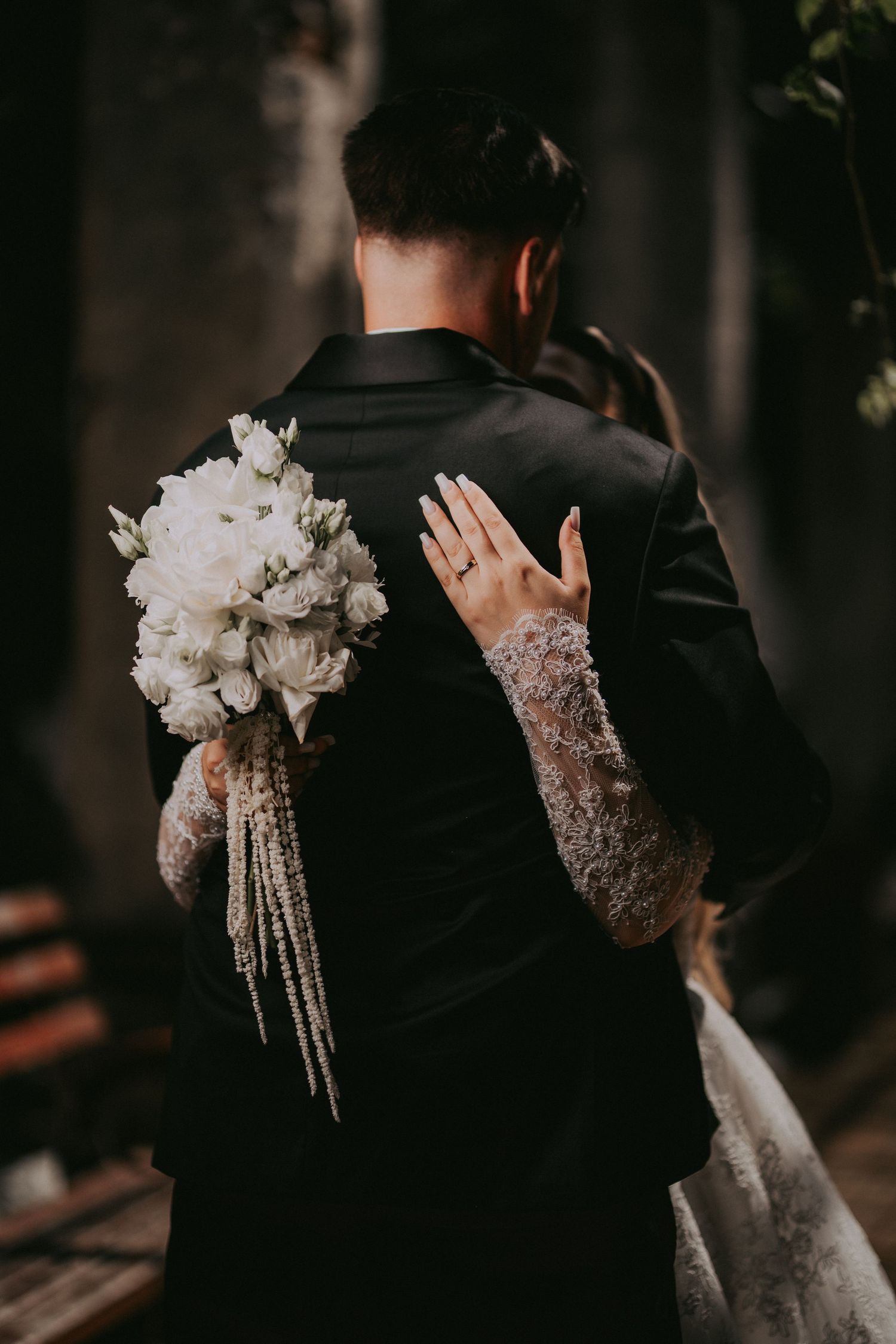 Bride embraces groom from behind holding a cascading white bouquet with elegant lace sleeves.