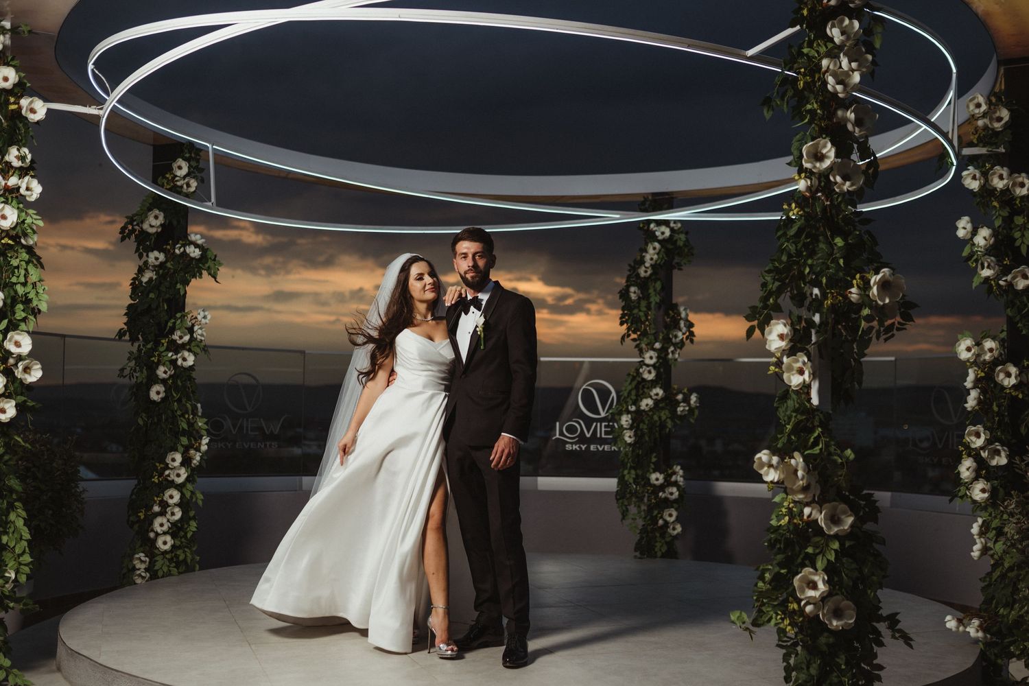 Bride and groom stand together under circular floral arch with neon lights at rooftop wedding venue at sunset.