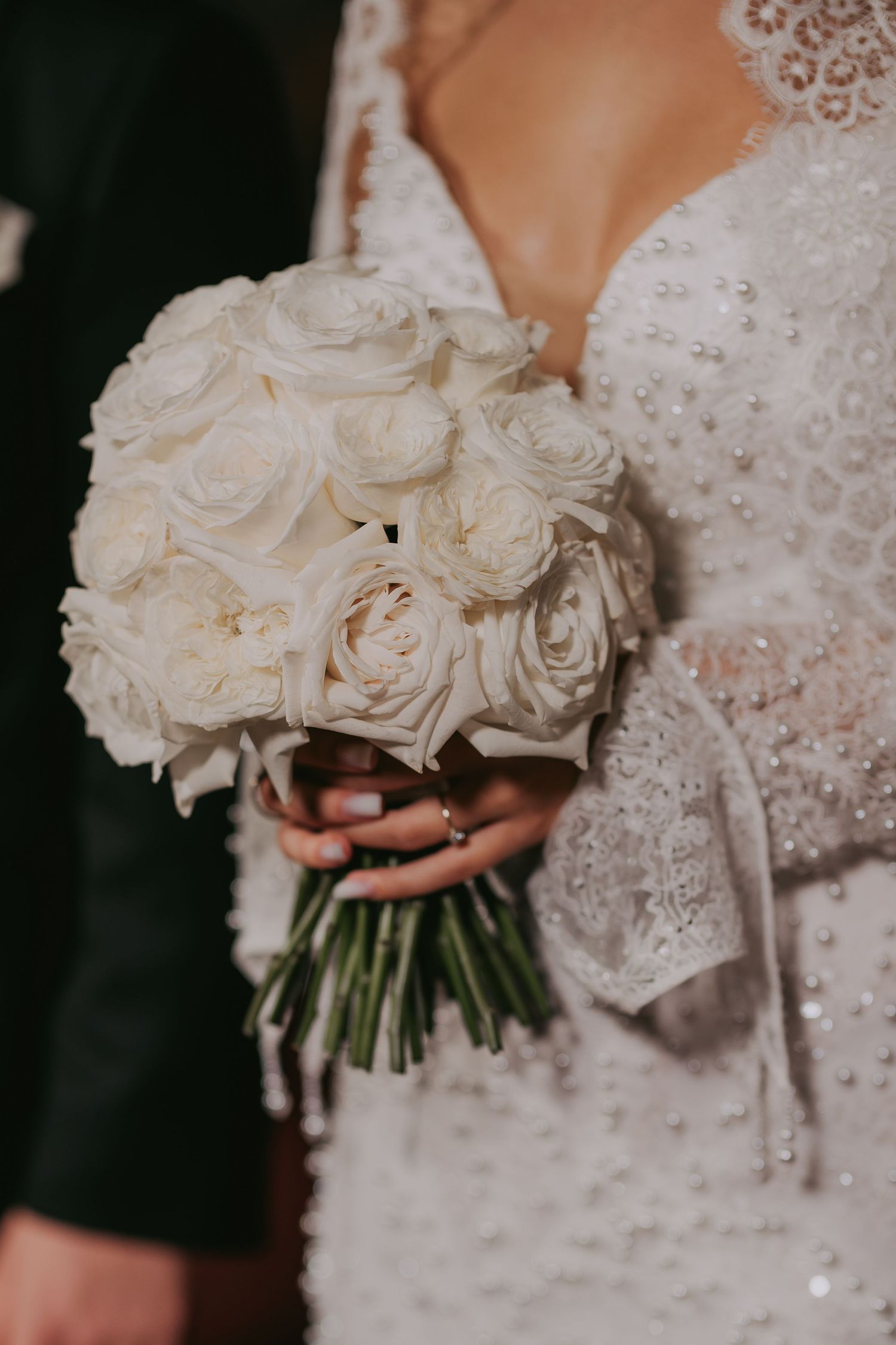 Bride holding elegant round bouquet of white roses with pearl-embellished lace wedding dress sleeves visible.