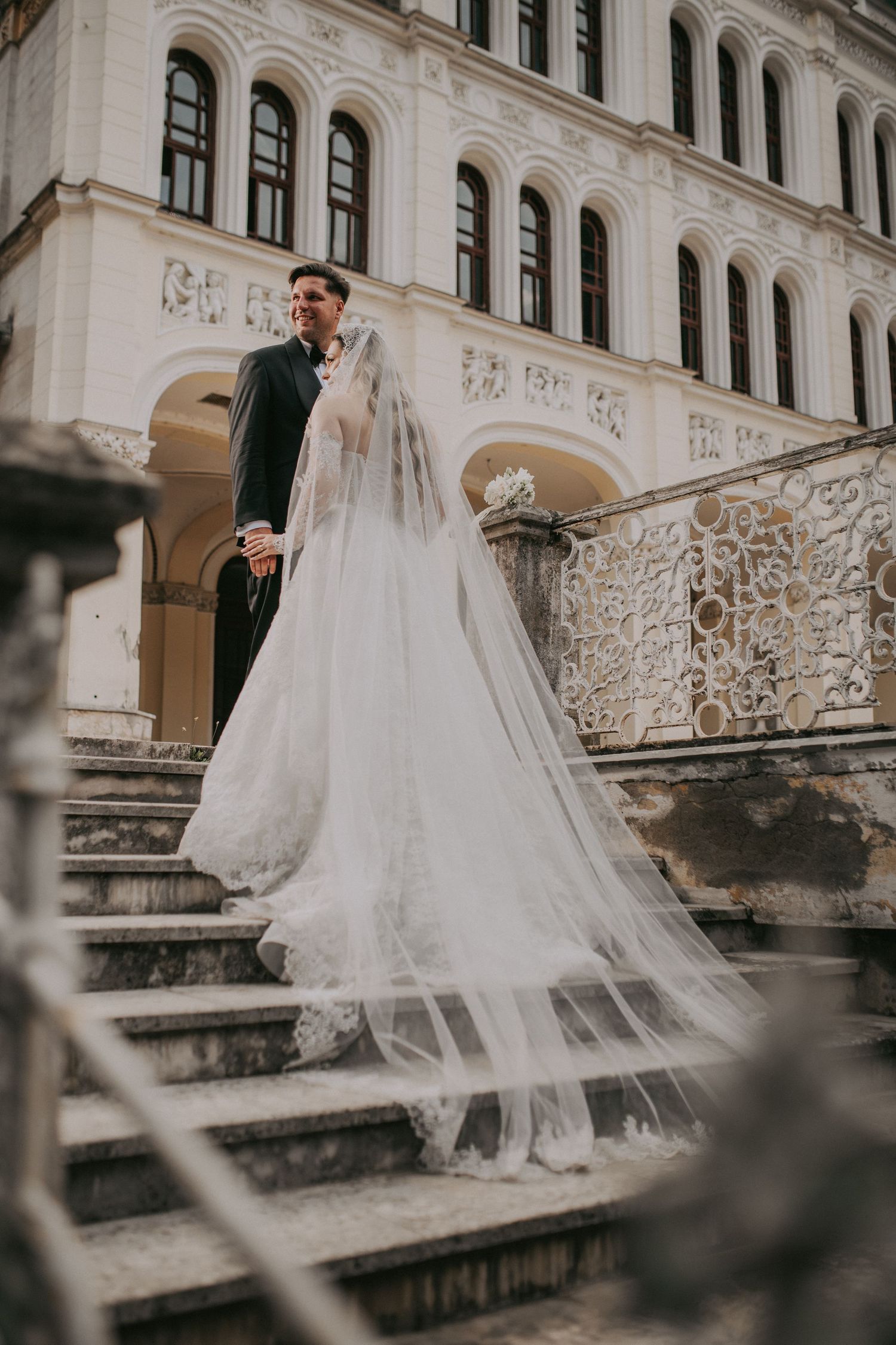 Bride and groom descend ornate stone steps of a grand historic building with flowing veil.