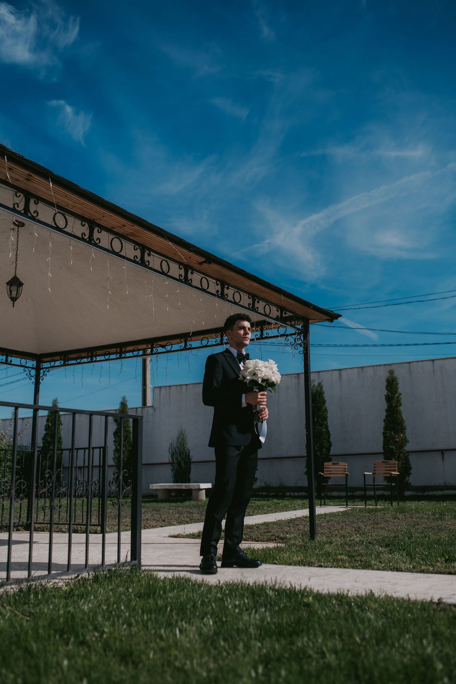 Groom in black suit holding a white bouquet standing under an outdoor gazebo on a sunny day. Epicu Fotograf Drobeta-Turnu Severin