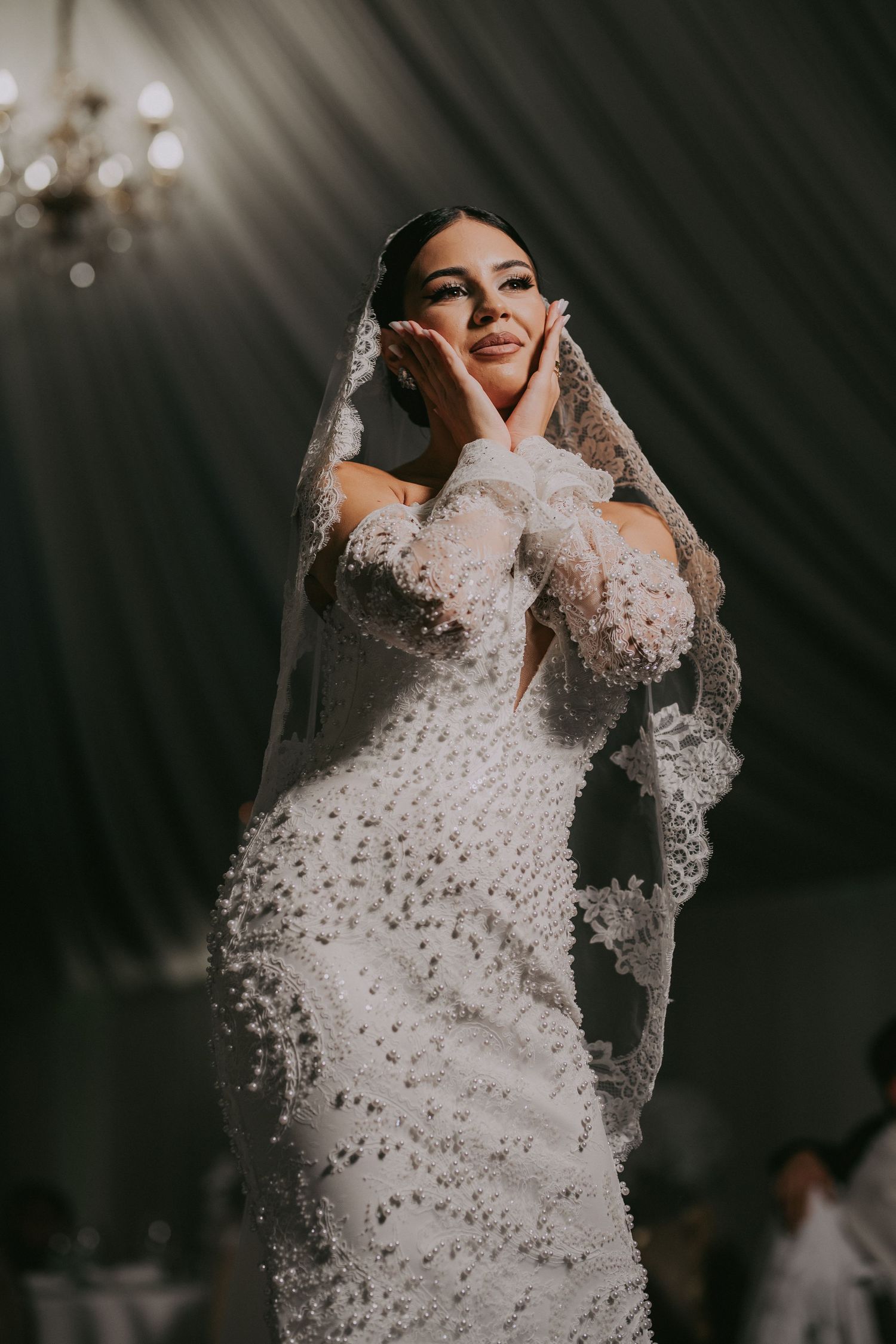 Bride in a pearl-embellished wedding gown and veil poses gracefully with hands framing her face under dramatic lighting.
