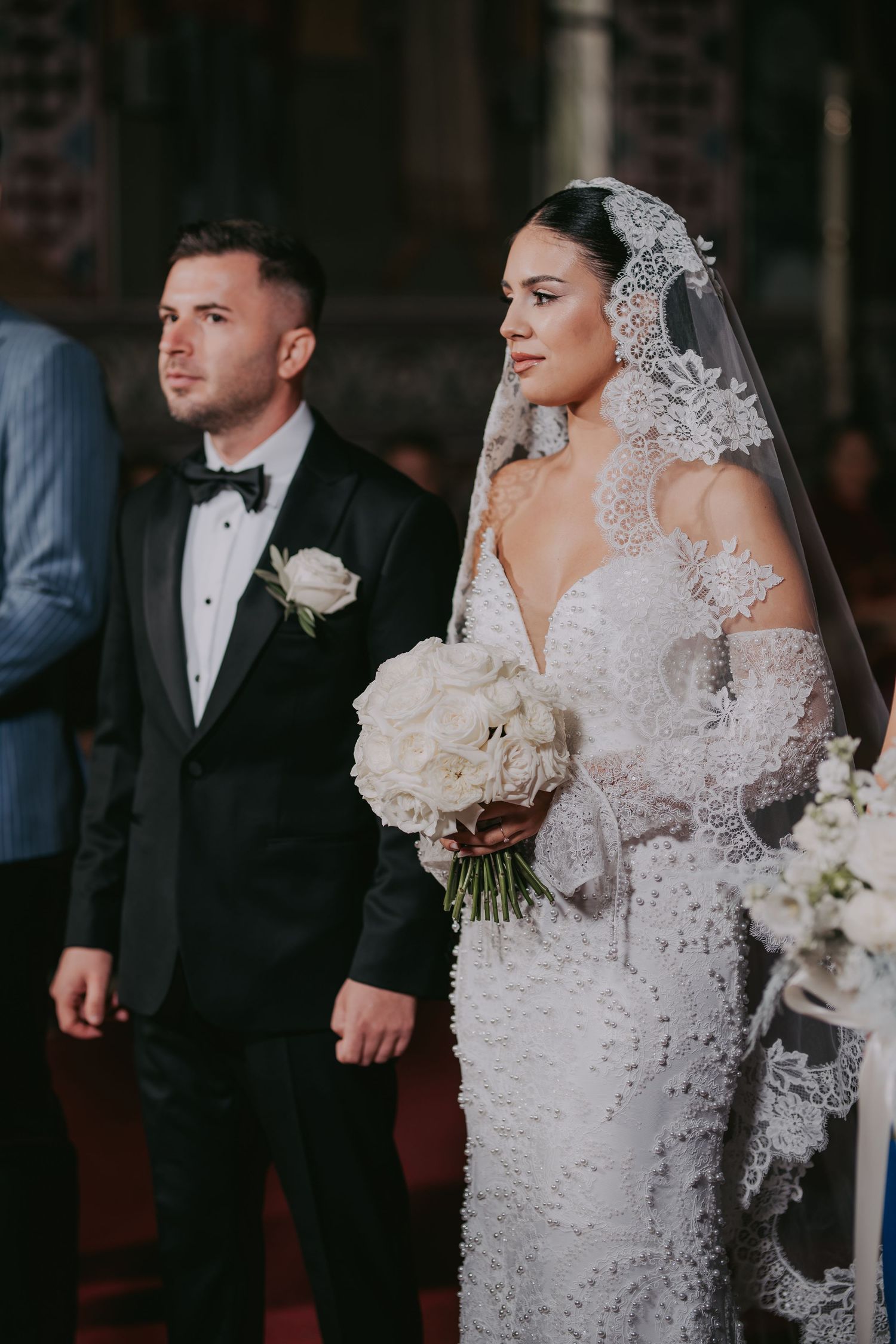Bride in lace veil and beaded gown walks with groom in tuxedo during church ceremony.