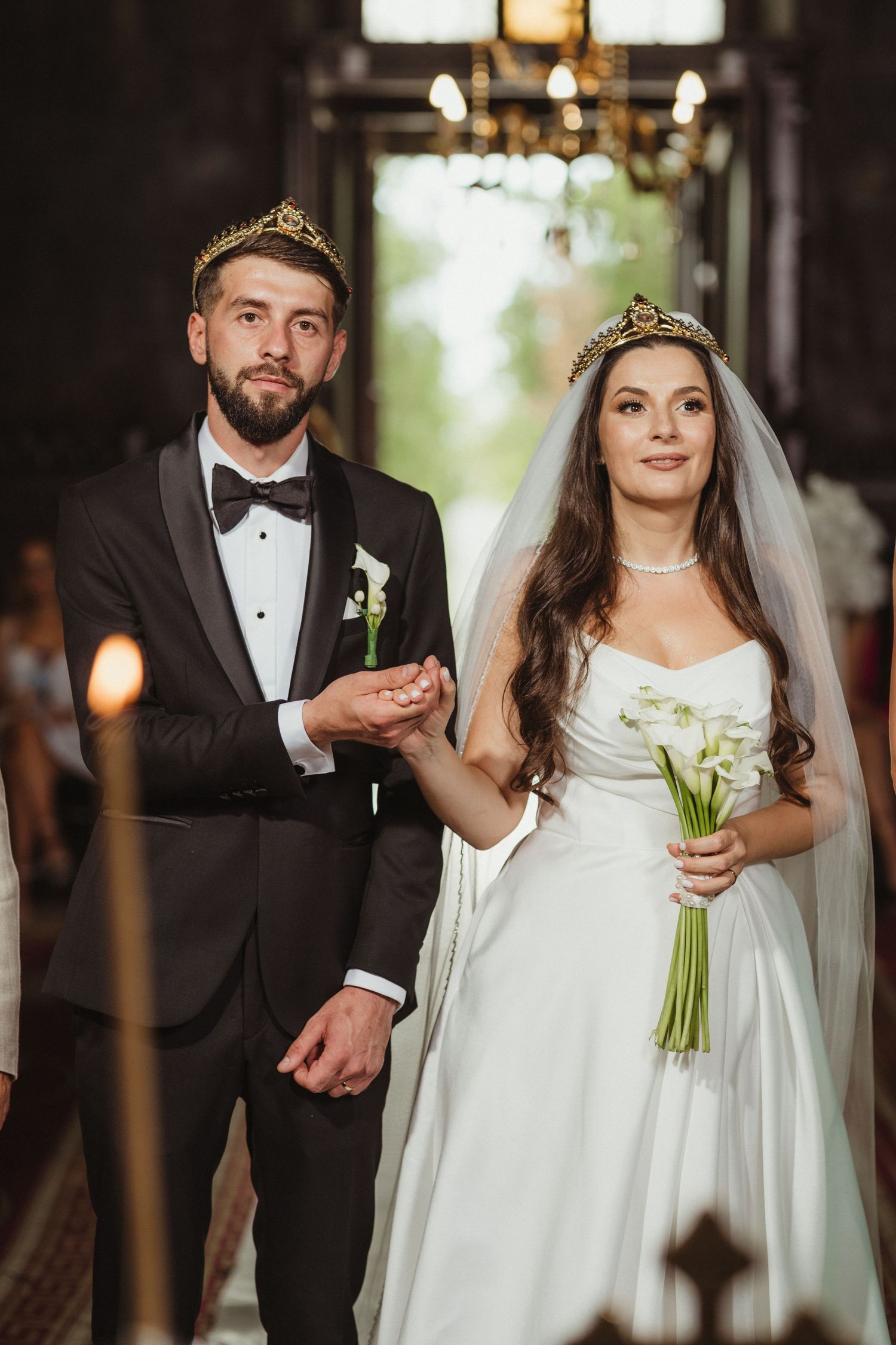 Bride and groom wearing golden crowns hold hands during Orthodox wedding ceremony inside an ornate church.