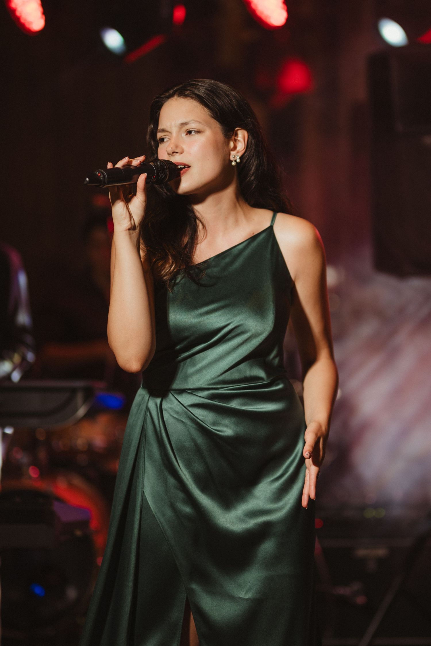 Woman in green satin dress singing into microphone on stage with red stage lighting.